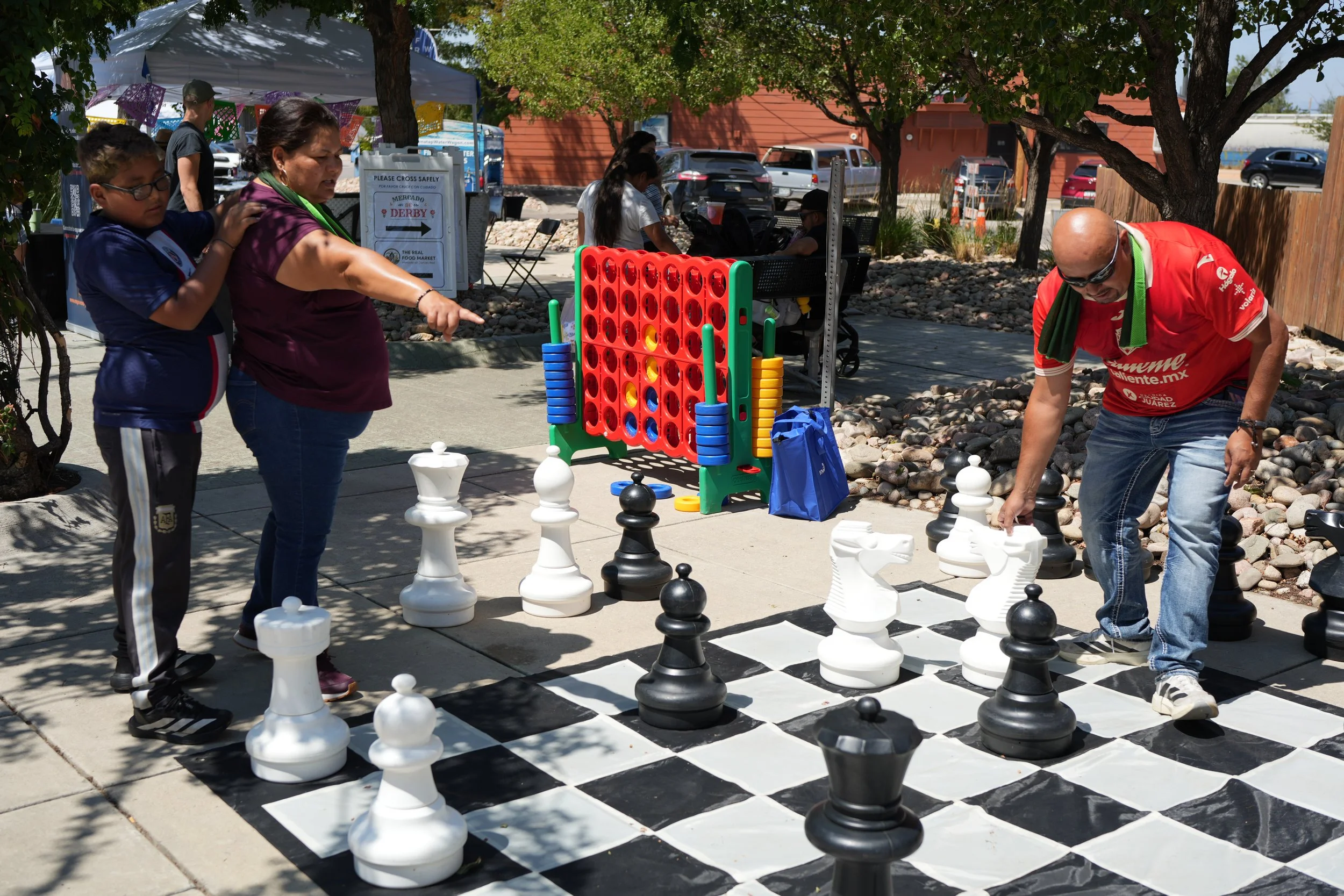 People playing giant chess outdoors under trees at a community event with tents and parked cars in the background.