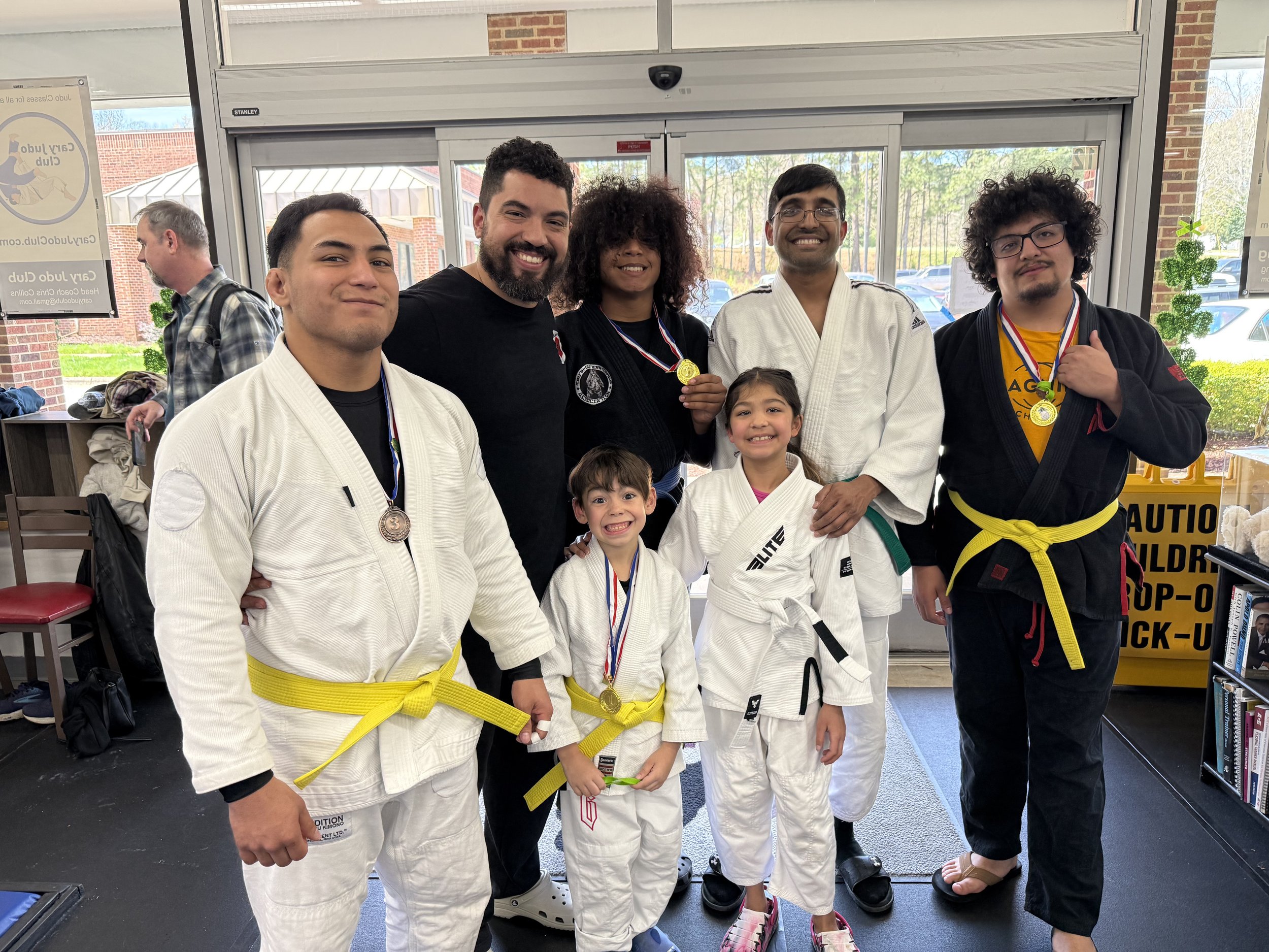 Group of five martial artists, three men and two young girls, smiling and posing inside a gym, wearing judo gis with medals around their necks, Judo, brazilian jiu jitsu, japanese jujutsu, jujitsu