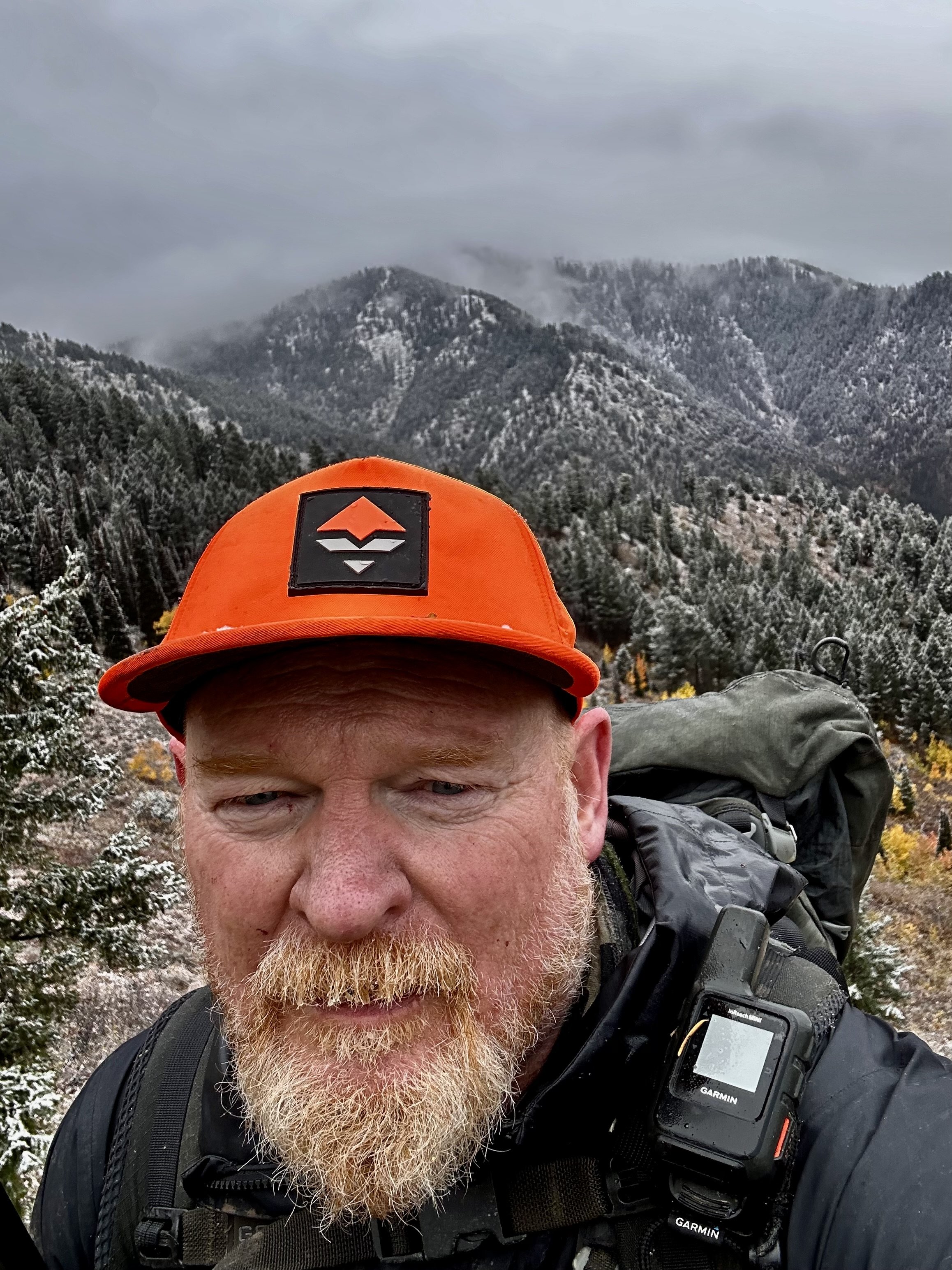 Close-up of a man with a white beard and reddish hair, wearing an orange cap, black jacket, and carrying a backpack and a Garmin device, with a mountainous landscape in the background.