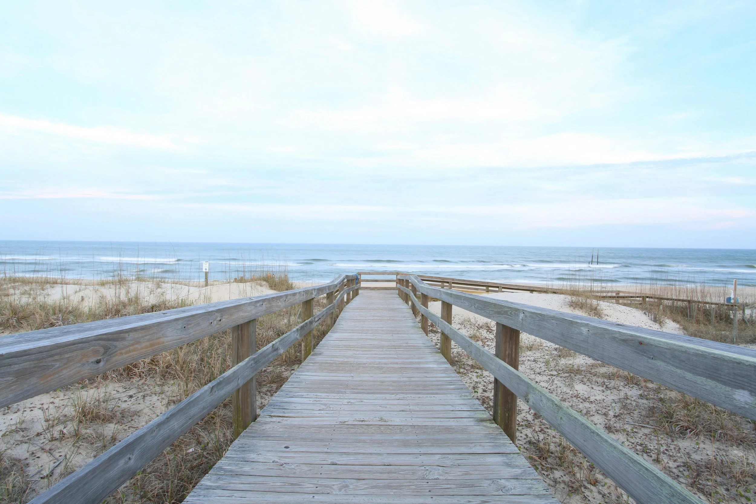 Wooden boardwalk leading to a beach with sand dunes, grasses, and the ocean in the background under a cloudy sky.