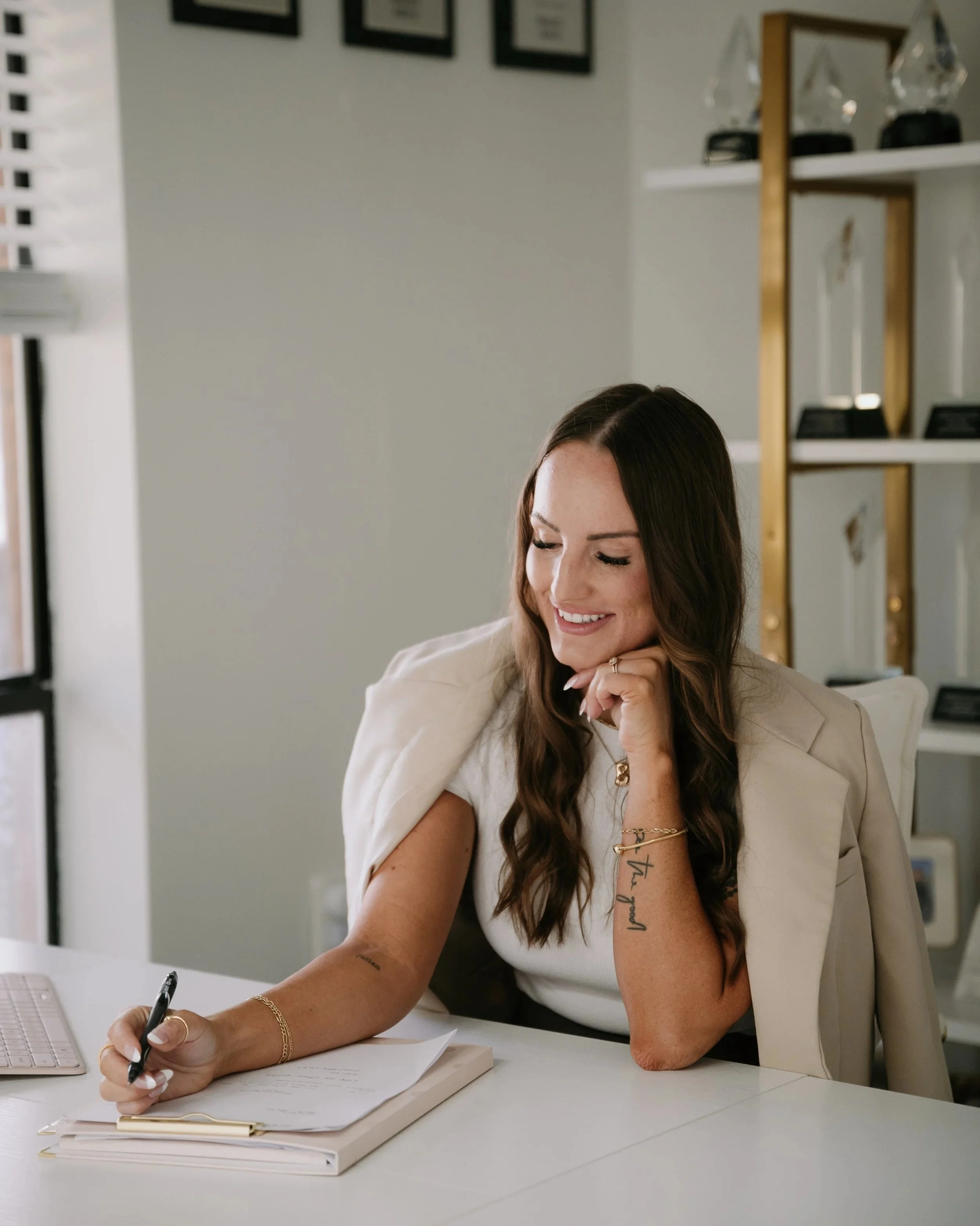 A young woman with long brown hair and tattoos on her arm sitting at a white desk, smiling and writing in a notebook with a black pen. She is wearing a beige blazer and gold jewelry. There are shelves with trophies and framed items on the wall behind her.