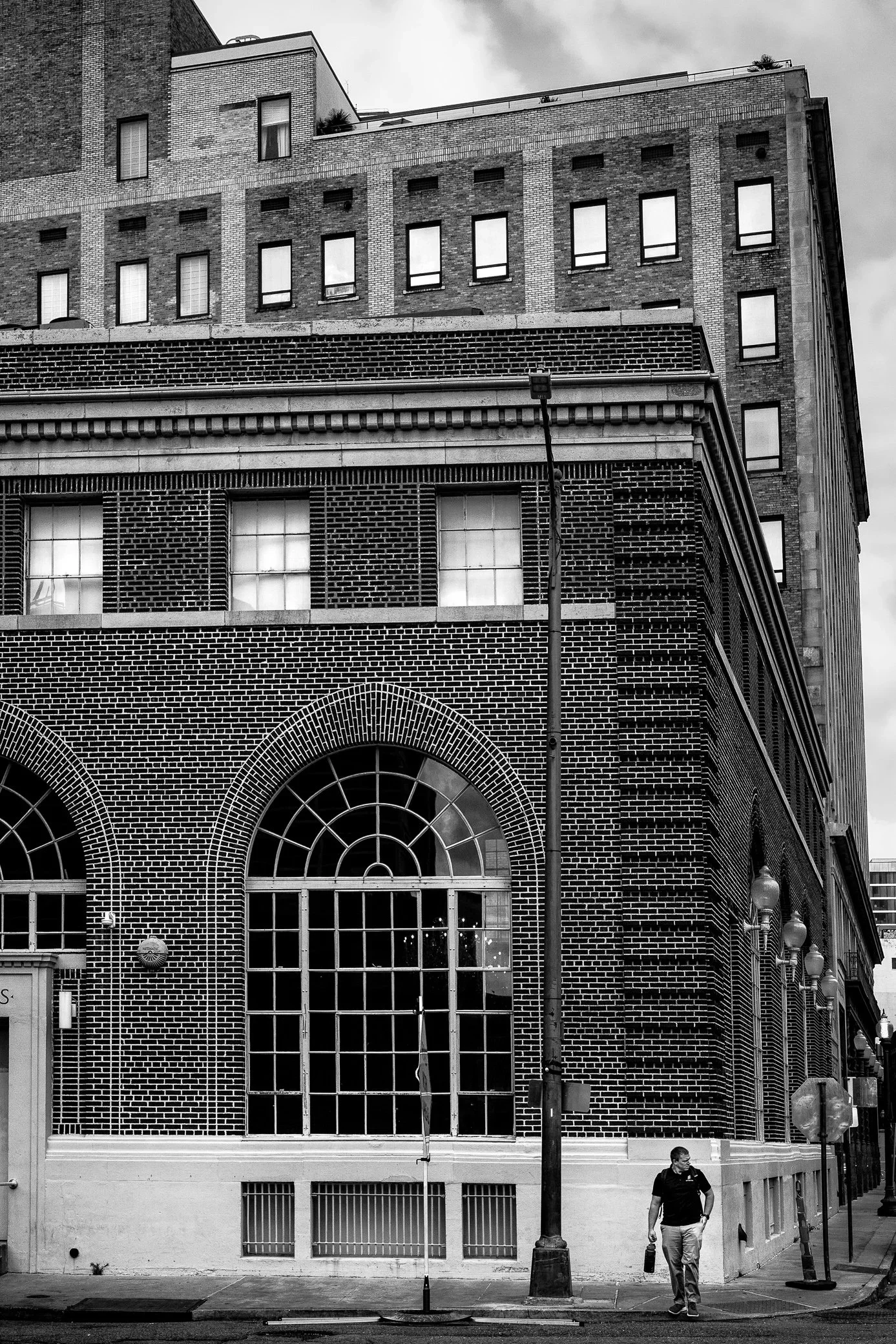 A lone man walks along the sidewalk at the foot of a tall brick building with arched windows, downtown New Orleans.