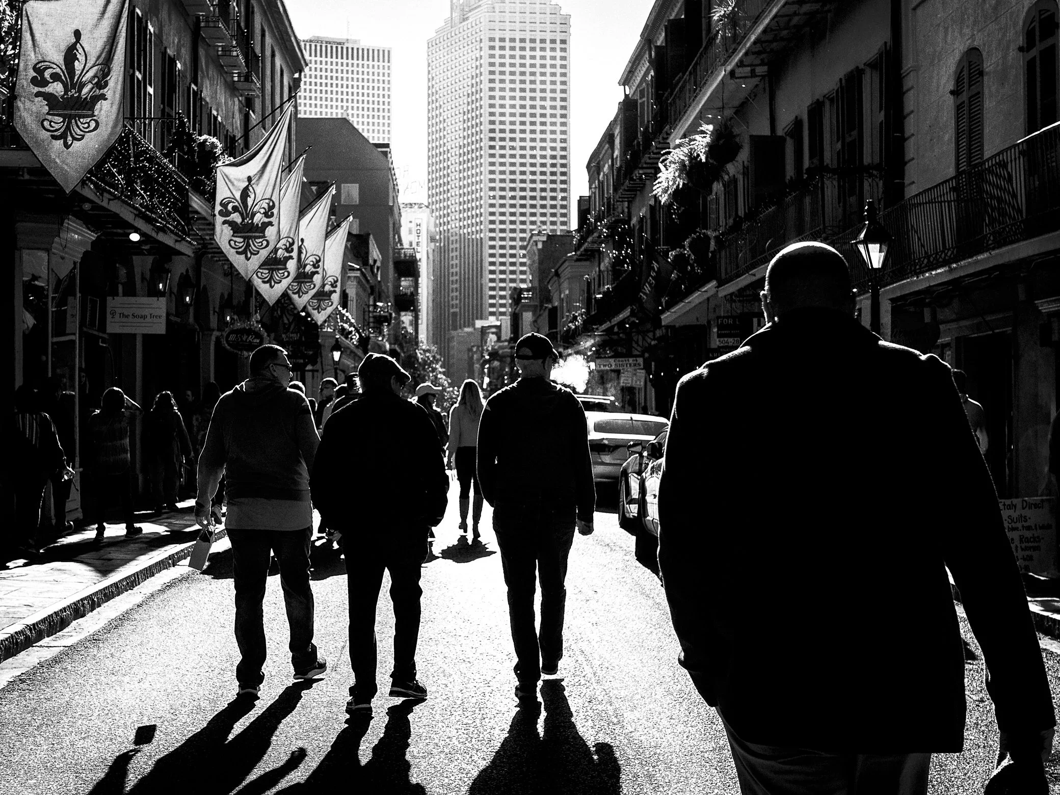 People walk in silhouette down a sunlit French Quarter street toward downtown towers, fleur-de-lis banners overhead and long shadows on the pavement.