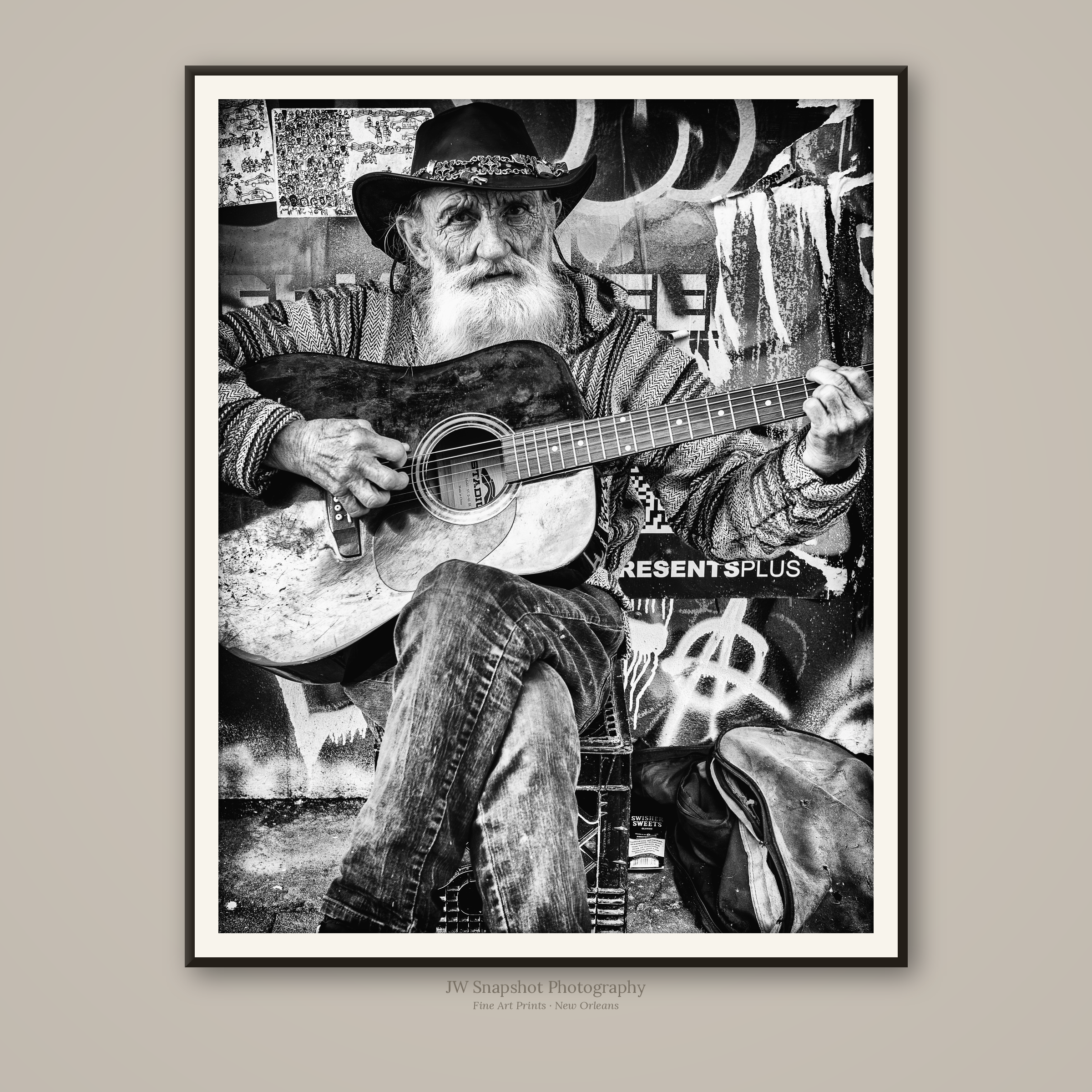 Black and white portrait of an elderly man with a long white beard playing an acoustic guitar, wearing a wide-brimmed black hat. Behind him are various artwork and graffiti-style decorations on the wall.