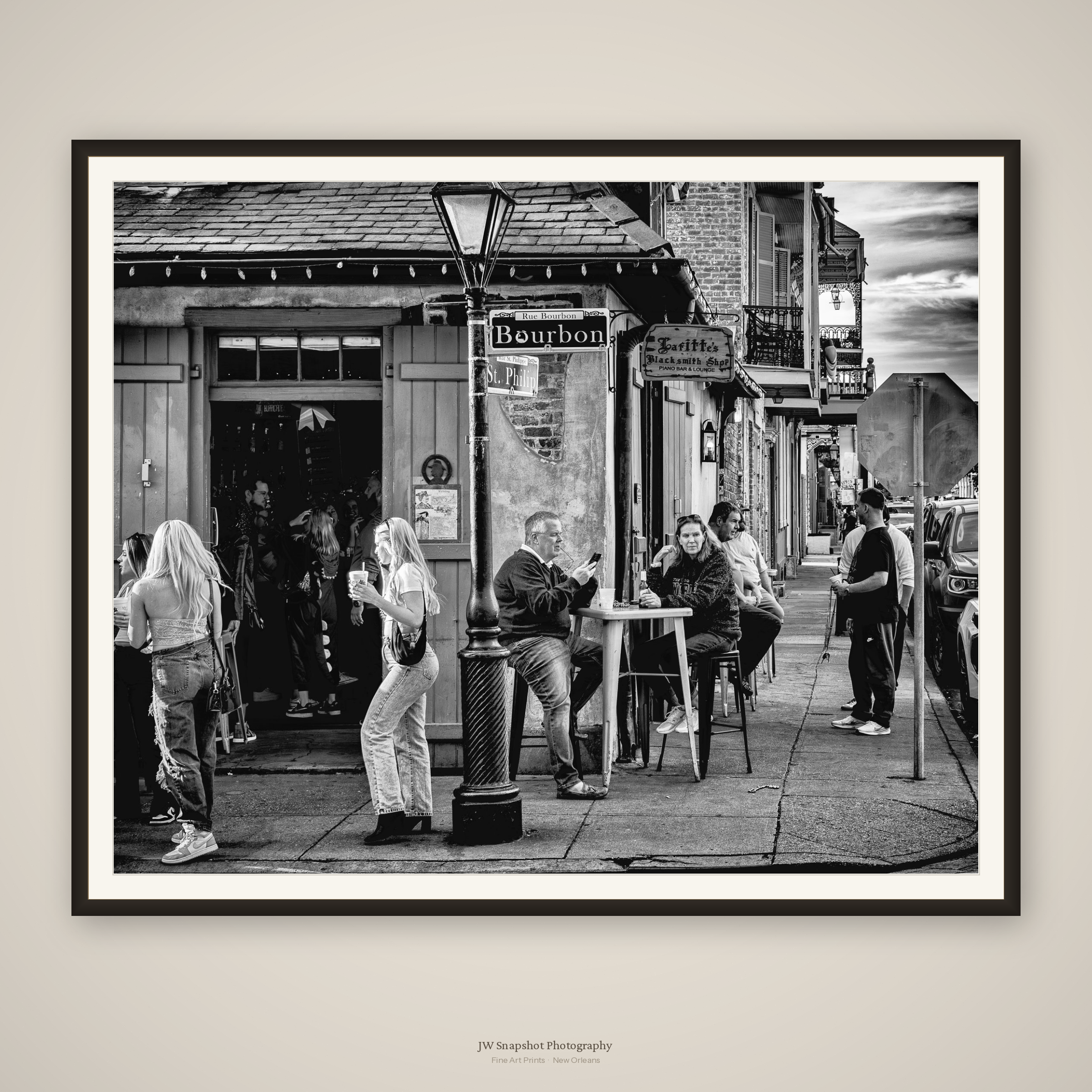 Black and white photo of a street scene on Rue Bourbon in New Orleans, showing patrons sitting at outdoor tables, a street lamp, and buildings with balconies and signage including 'Taffy's Blacksmith Shop'.