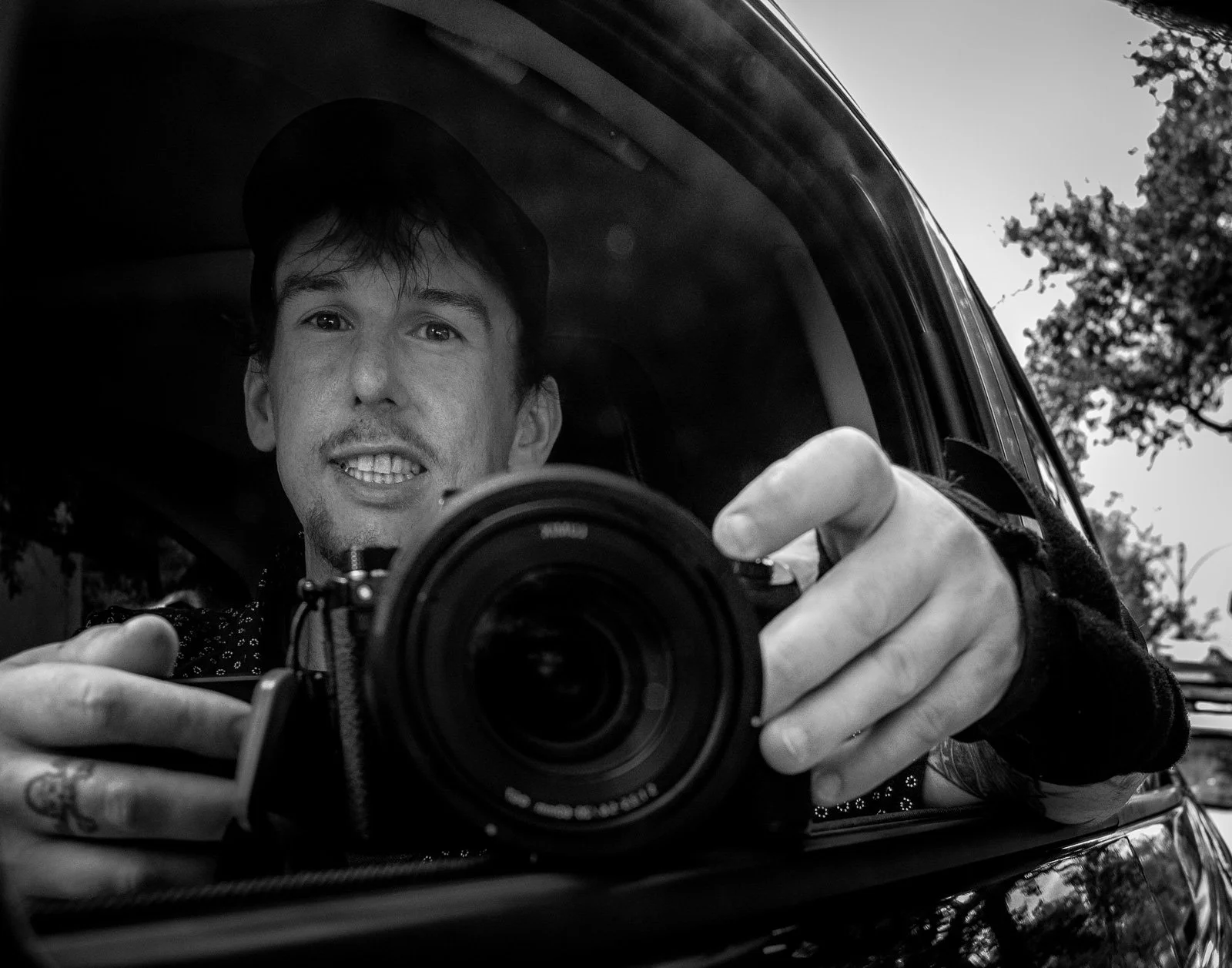 A man taking a selfie with a camera while looking into a car's side mirror. The photo is in black and white, and the man is wearing a cap.