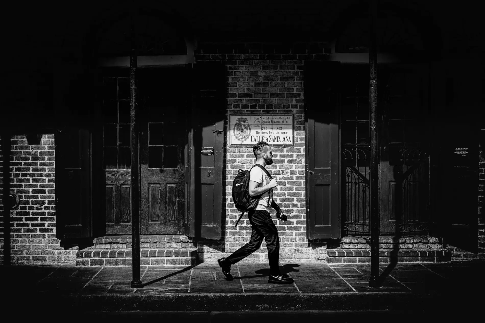 A man with a backpack walks through a band of hard sunlight past a brick wall and the Calle de Santa Ana street plaque.