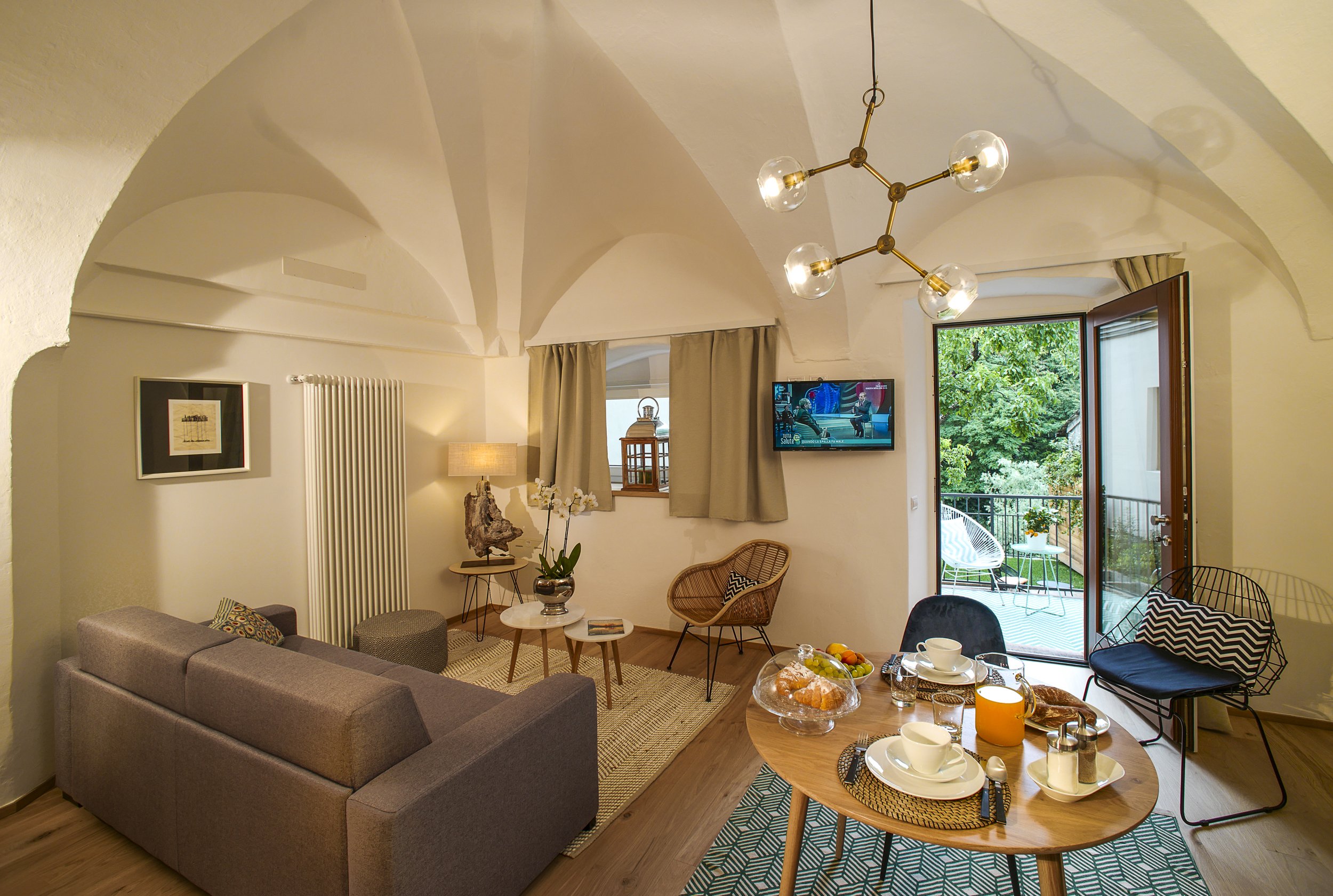 A cozy living room with a gray sofa, a wooden coffee table with breakfast dishes, a black wire chair, a round side table with a decorative sculpture, a television, and a large open door leading to a balcony with outdoor furniture. The room has beige 
