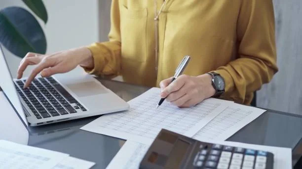 Person in yellow shirt working with a laptop, paperwork, and calculator on a desk.