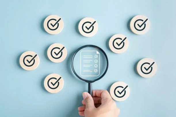 Hand holding a magnifying glass over a checklist surrounded by several other checkmarks on round tokens on a blue background.