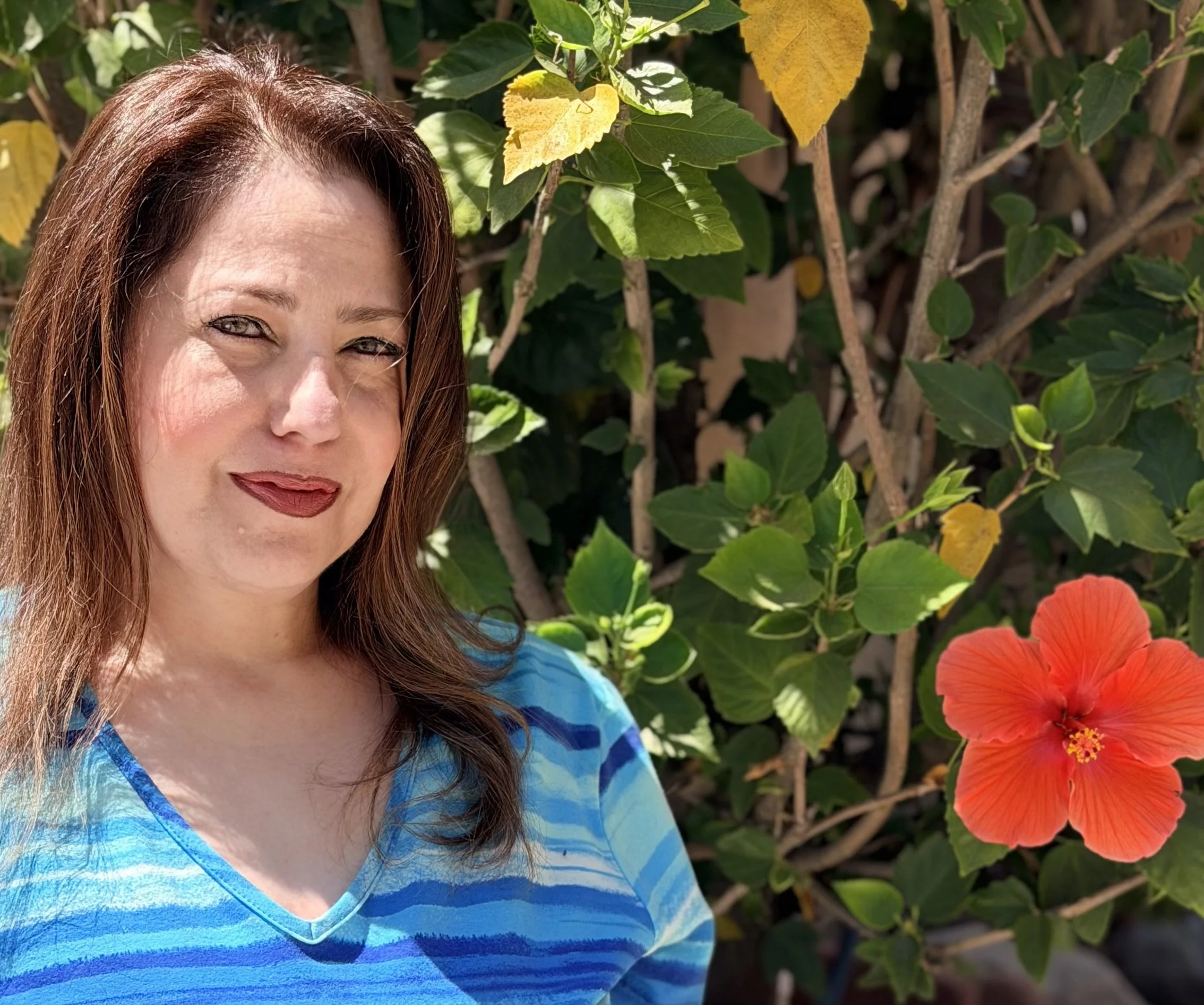 A woman with long reddish-brown hair and light-colored eyes, wearing a blue tie-dye shirt, stands outdoors in front of green and some yellow leaves, with a bright orange hibiscus flower visible in the bottom right corner.