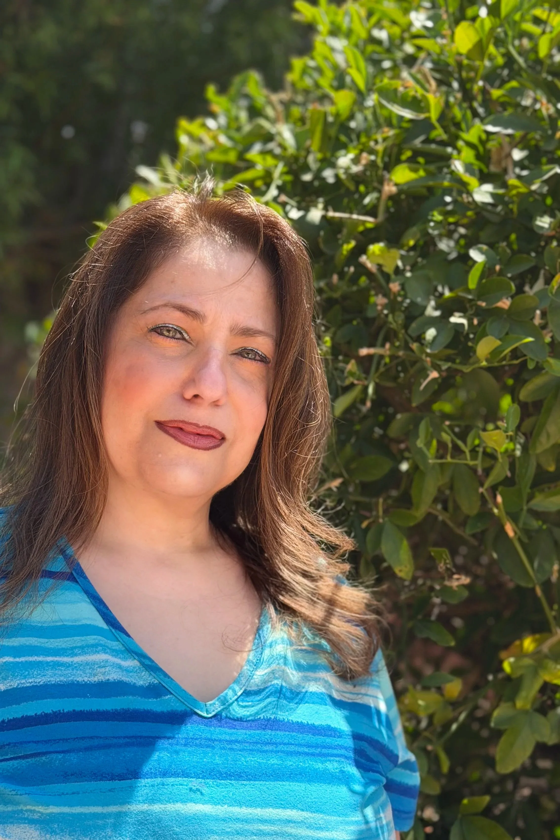 A woman with long brown hair and light skin smiling outdoors, standing in front of green leafy bushes, wearing a blue striped V-neck shirt.