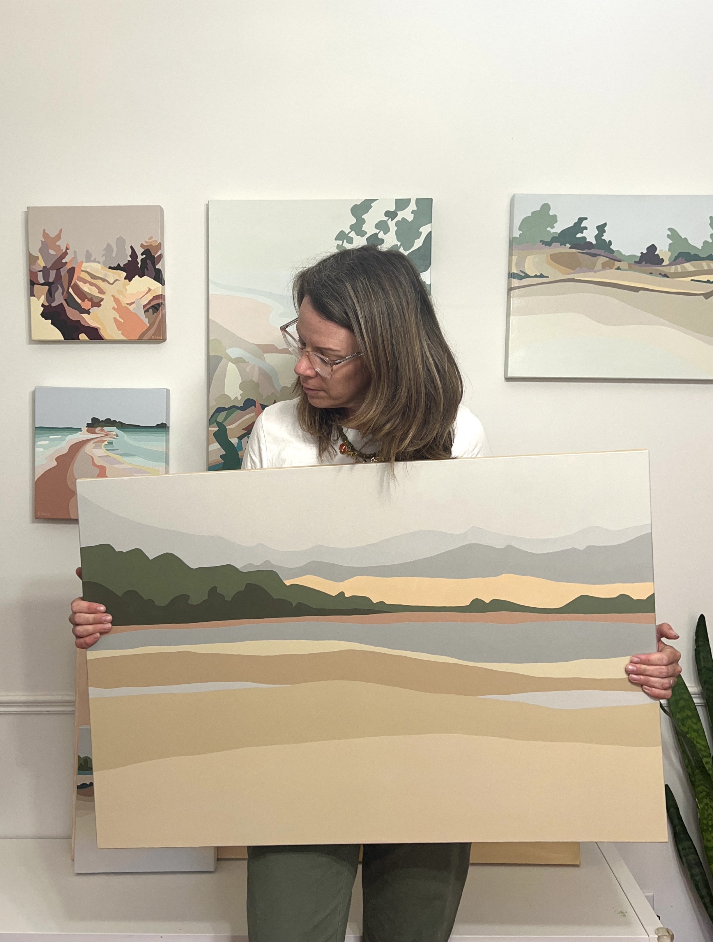 A woman wearing glasses holding a landscape painting in front of her, with similar landscape paintings on the wall behind her.