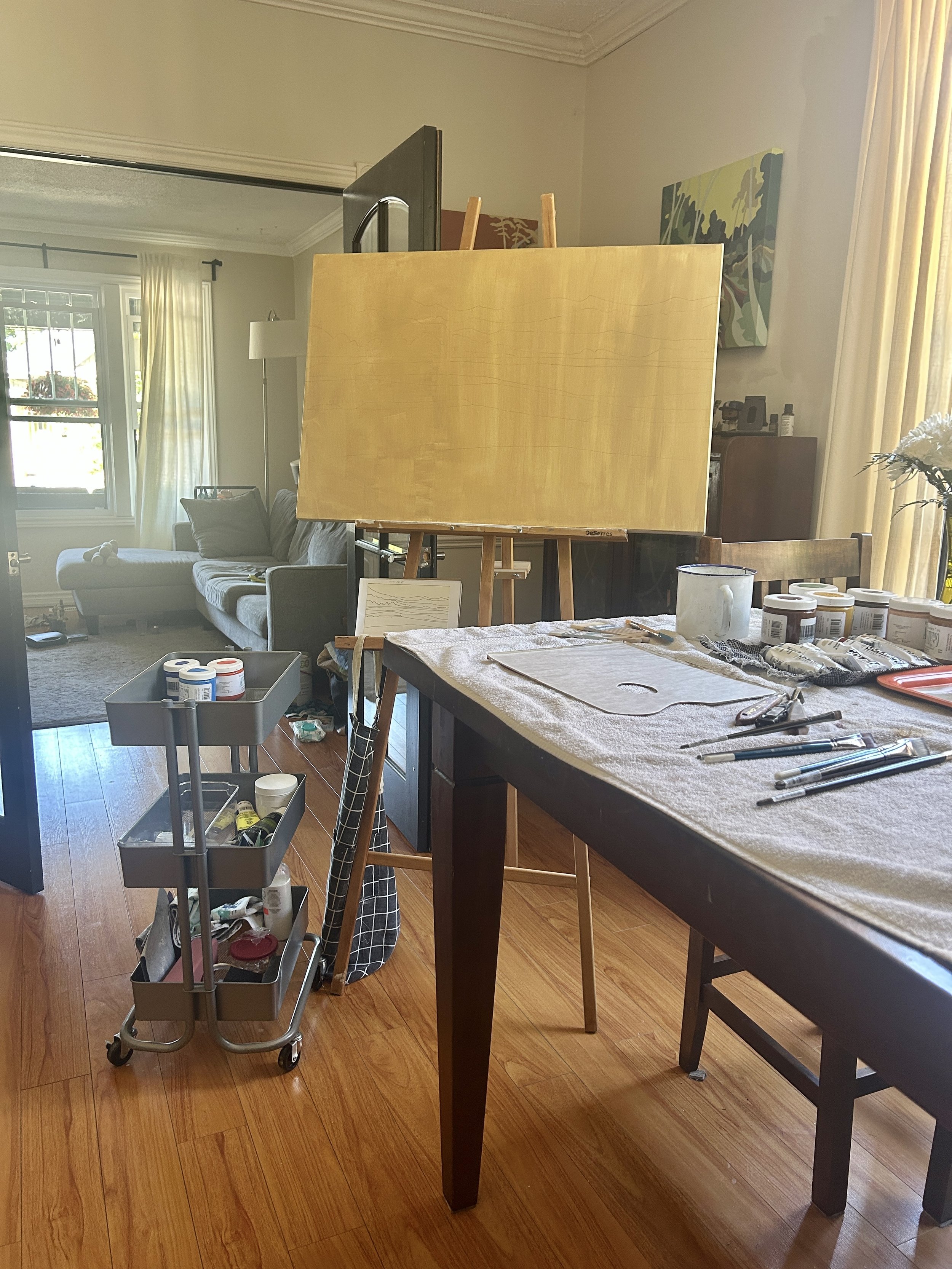 An artist's workspace in a living room with a large canvas on a stand, painting supplies on a table, and a rolling cart filled with paints. There is a sofa with a pillow and a lamp in the background, with windows and curtains letting in natural light.
