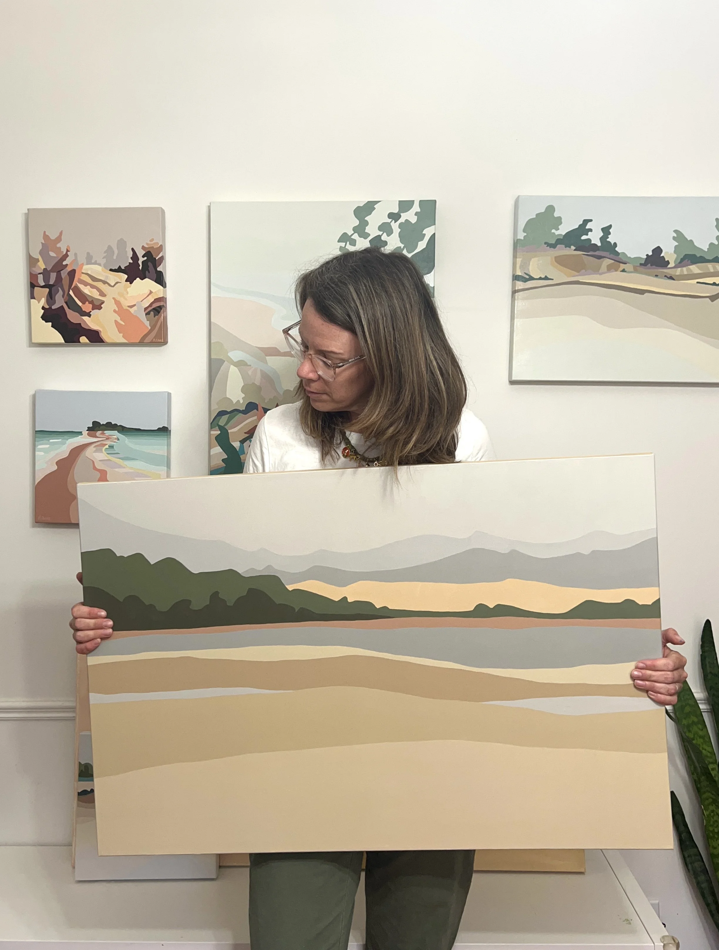 A woman holding a landscape painting with layered hills and sky, standing in an art gallery with additional paintings on the wall behind her.