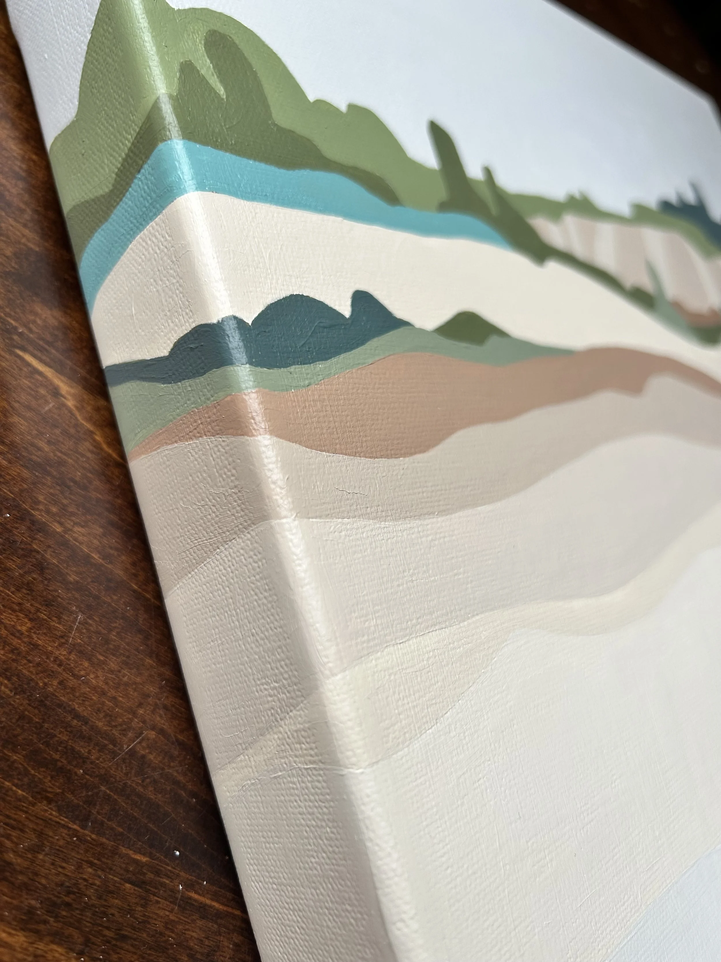Close-up of a canvas painting featuring abstract landscape with green hills, blue water, and beige land, placed on a wooden surface.