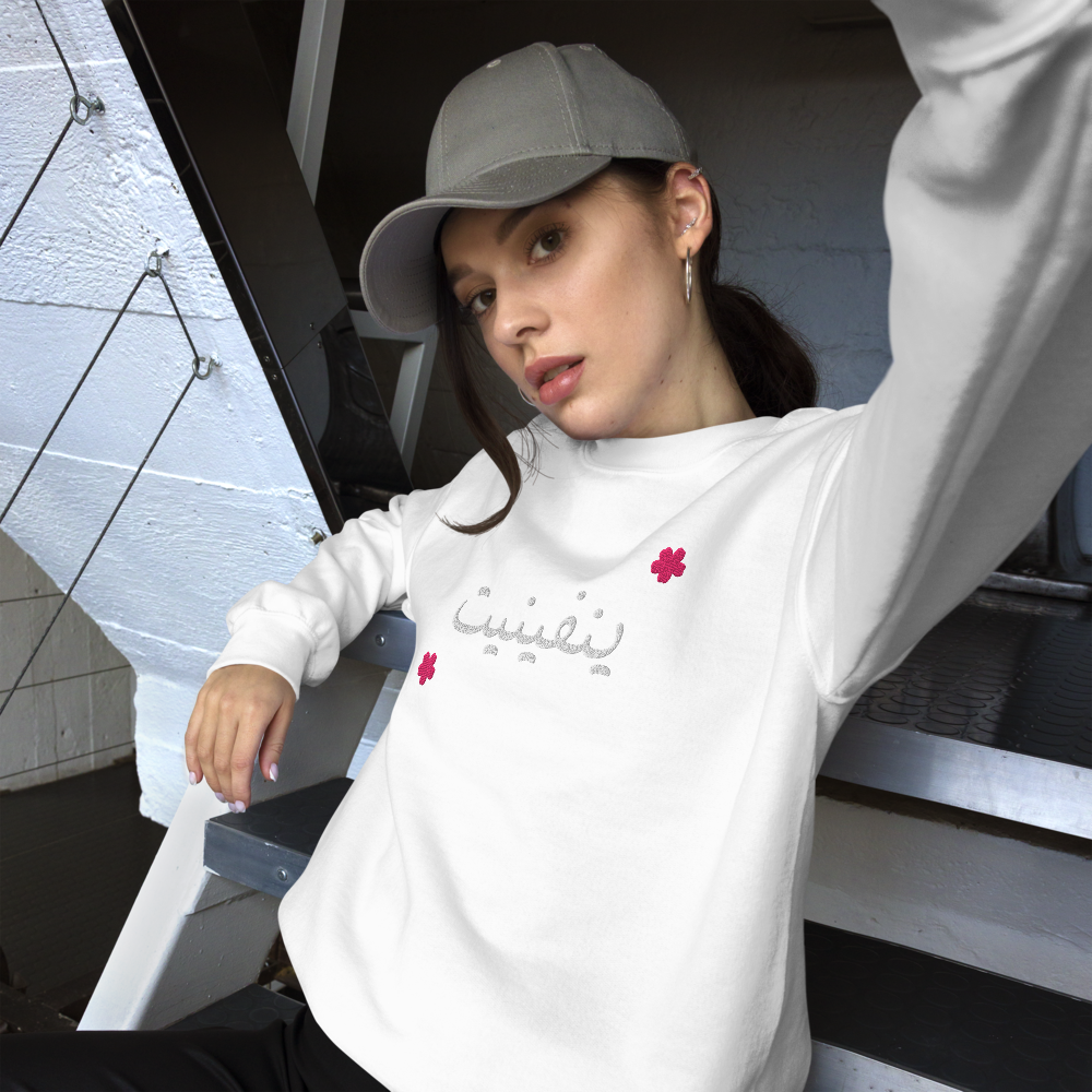 Jeune femme portant une casquette grise, une chemise blanche avec des fleurs roses et une écriture en arabe, assise sur un escalier métallique, regardant l'appareil photo.