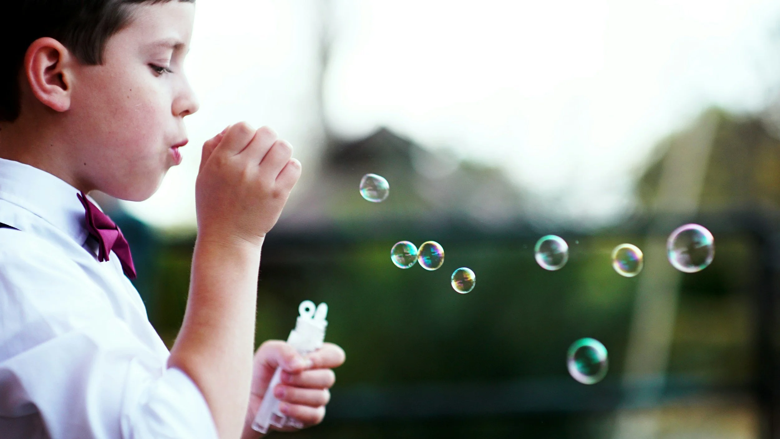Young boy in a white shirt and maroon bowtie blowing bubbles outdoors.