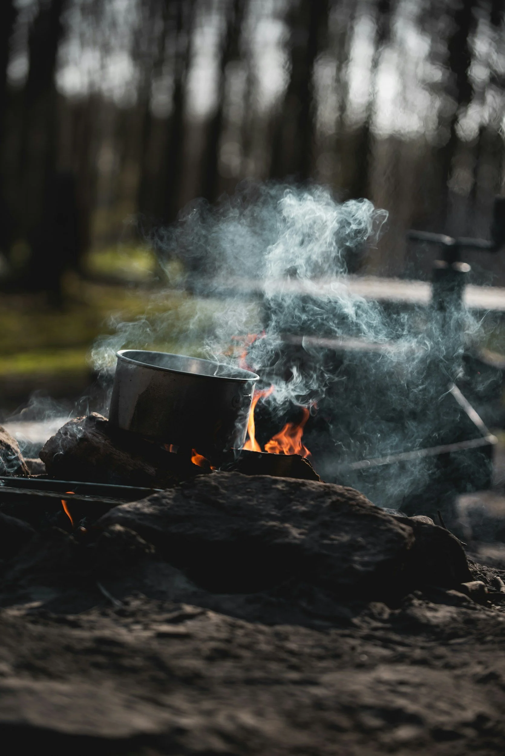 A metal cup placed over a campfire with flames and smoke, outdoors with trees in the background.