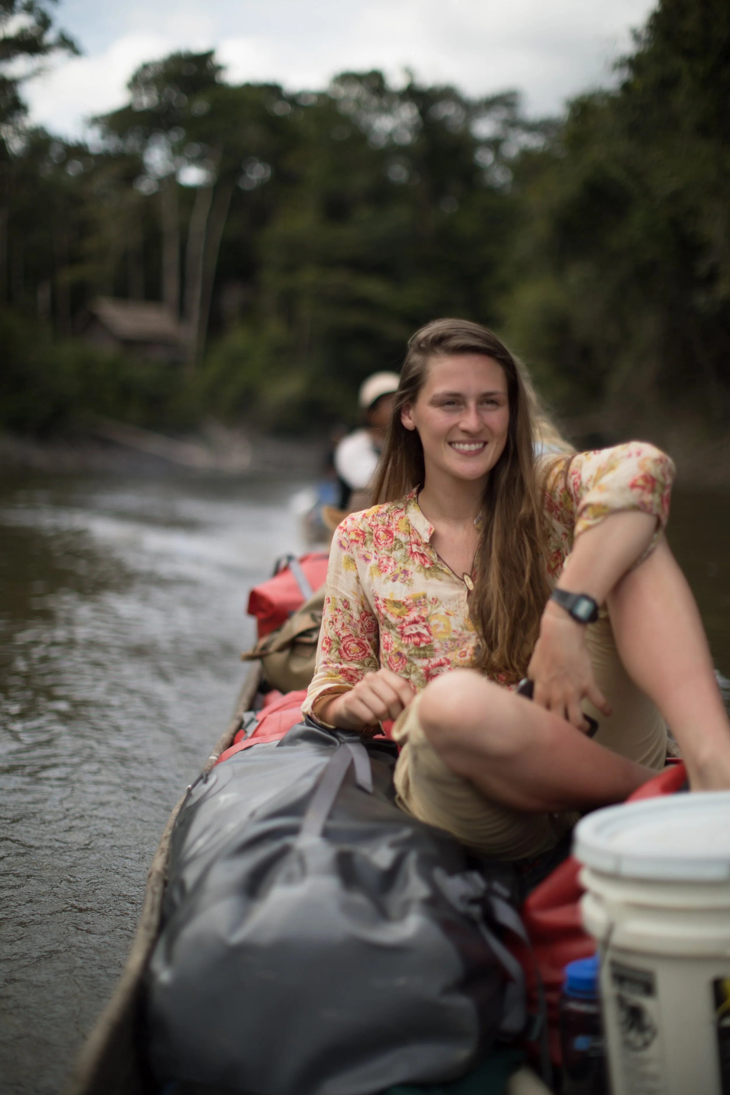Smiling woman in a floral shirt sitting on a boat with luggage and supplies, paddleboarding on a river surrounded by trees and a cloudy sky.
