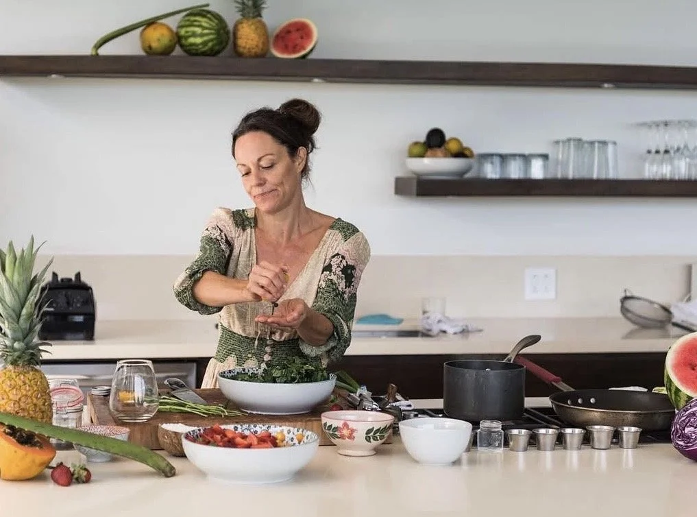 Woman preparing food in a modern kitchen with fresh fruit including pineapple, watermelon, and papaya, and various bowls and utensils on the counter.