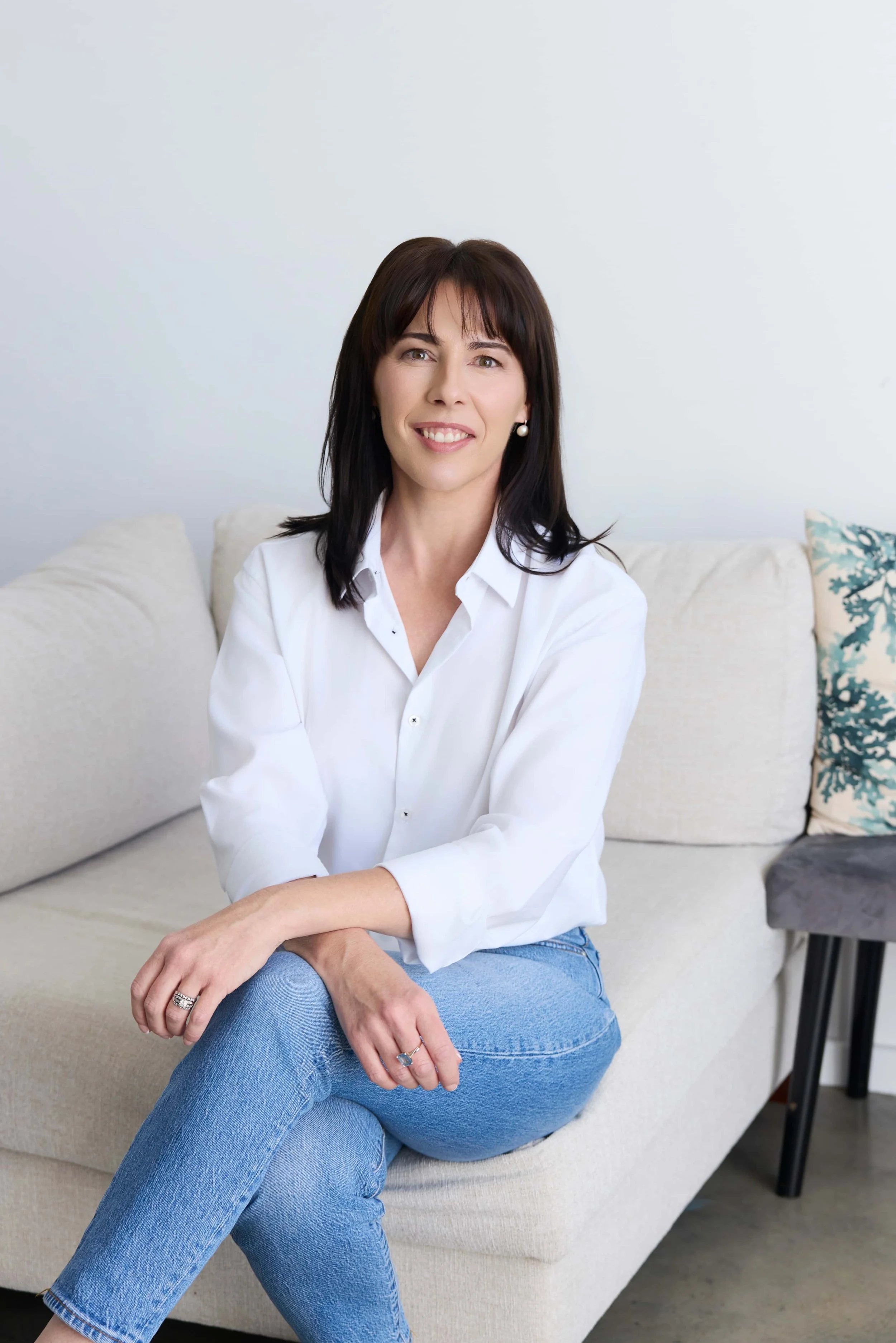 Lawyer, Taegan Irving, wearing a white shirt and blue jeans, sitting on a light-coloured couch with cushions, smiling at the camera.