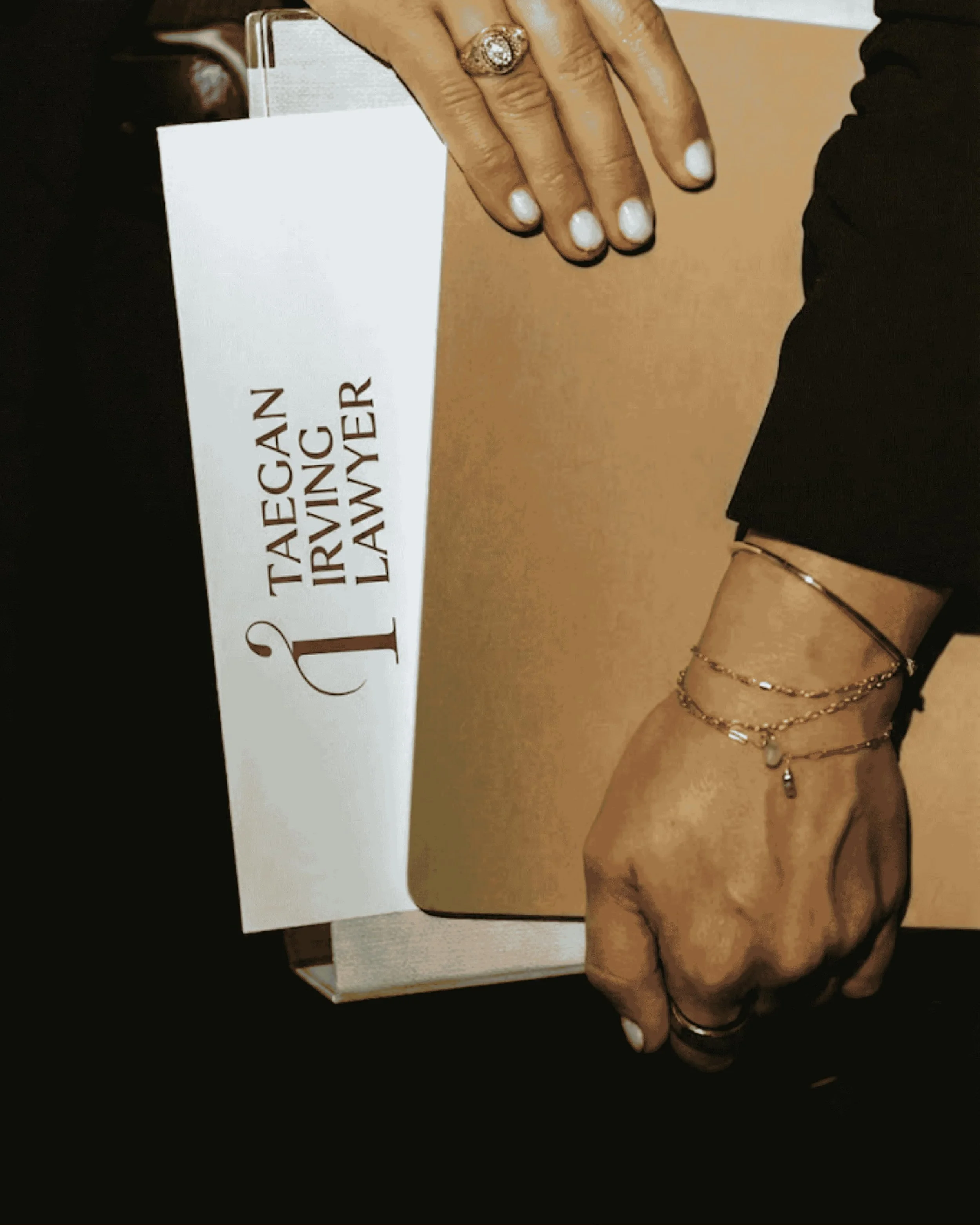 Close-up of a person’s hand resting on a surface, wearing rings, with multiple bracelets on the wrist, and a partial view of their outfit which includes black clothing and accessories. A sign next to the hand has partially visible text that reads, 'TAEGA... IRVING...AWER'.