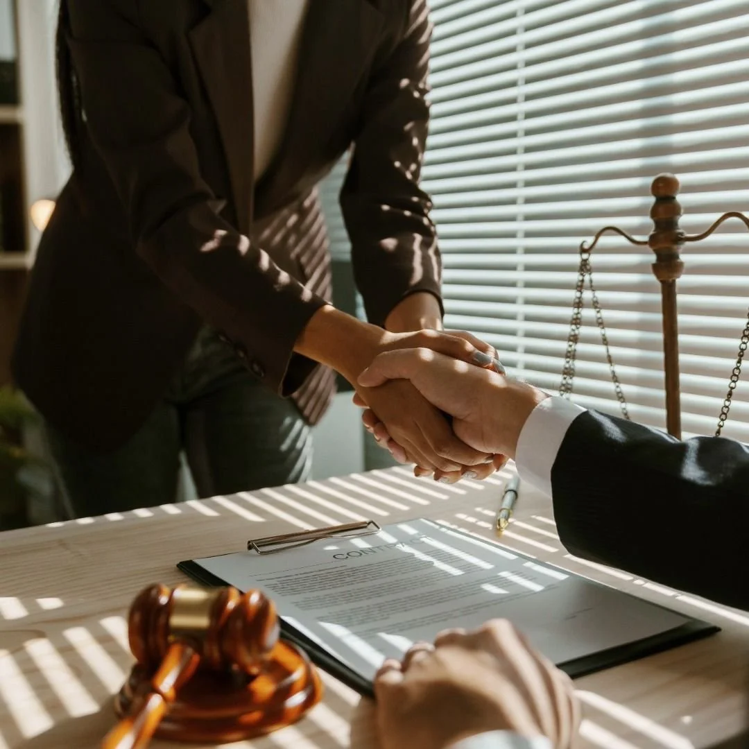 A person in a dark blazer shaking hands with another person in a suit over a desk during a formal agreement or contract signing, with legal symbols and documents visible.