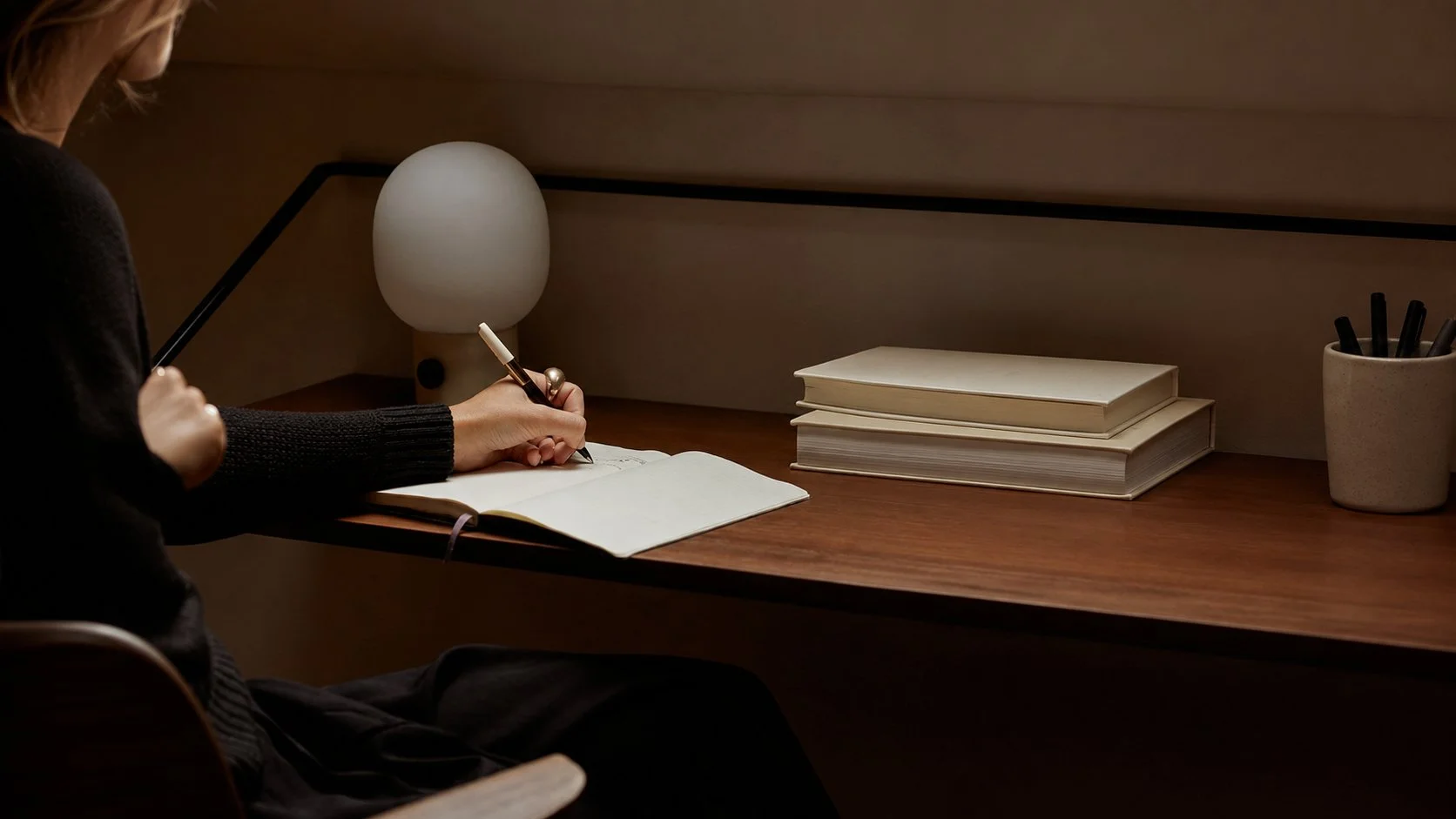 A person writing in a notebook at a desk with a modern lamp, a stack of white books, and a cup filled with black pens.