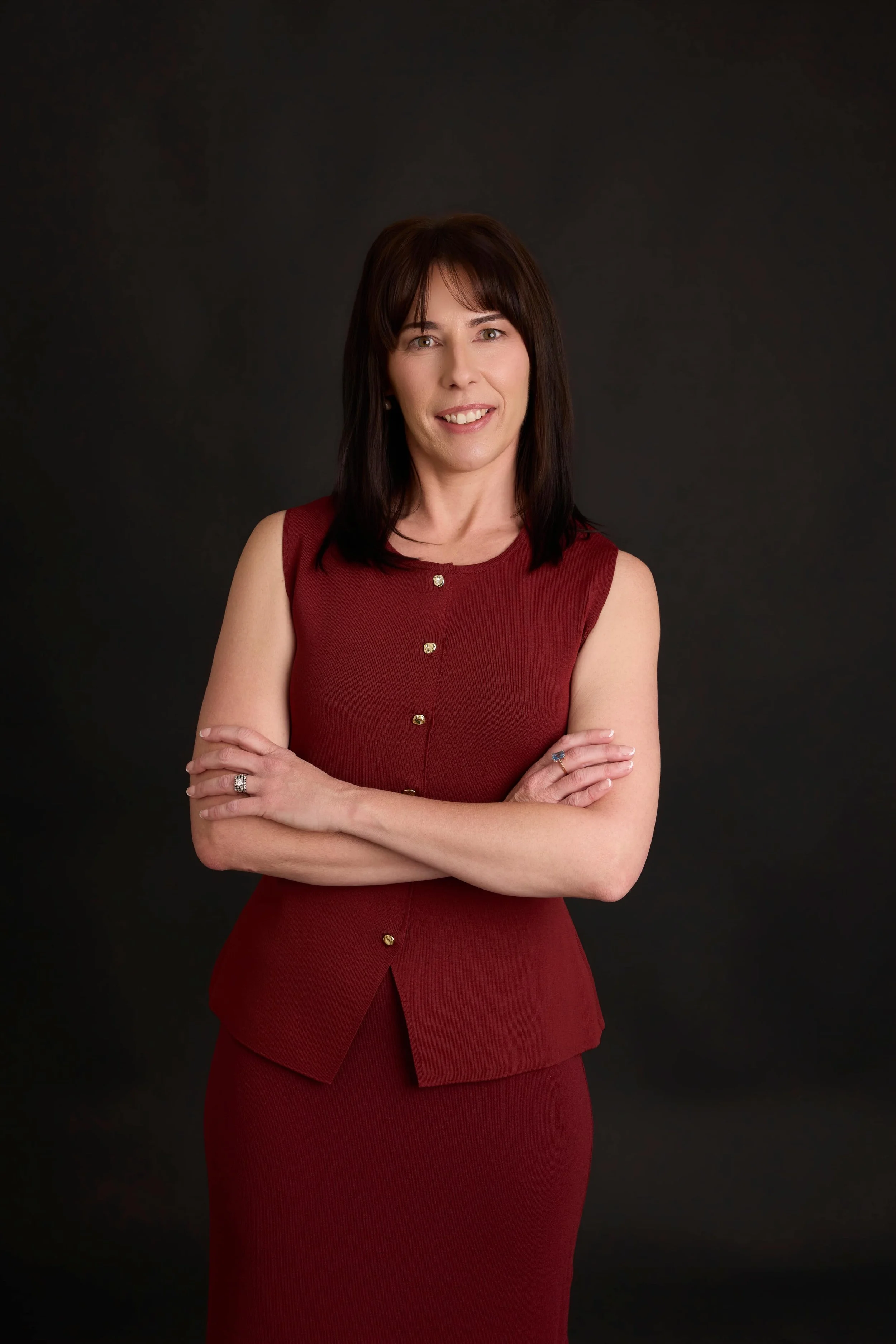 Lawyer, Taegan Irving, with dark brown hair wearing a sleeveless red dress with gold buttons, standing with arms crossed against a black background.