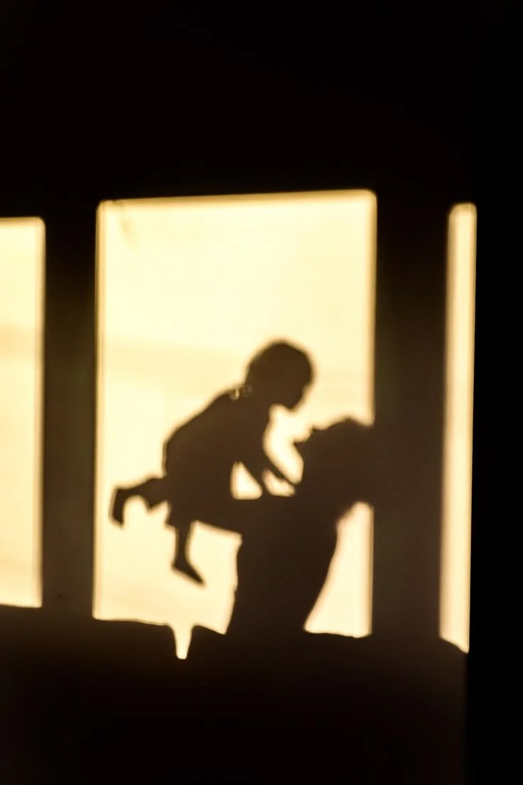 Silhouette of a child sitting on an adult's shoulders, in front of bright window light.