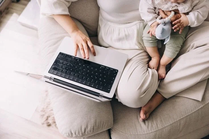 A person sitting on a beige couch with a laptop on their lap and a young child sitting beside them, holding a toy.