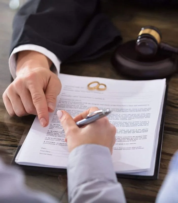A legal professional, identifiable by a black suit and white shirt, points to a document on a wooden table while the client, in a light shirt, signs it with a pen. Two gold wedding rings are on the document, and a gavel with a sound block is in the background.