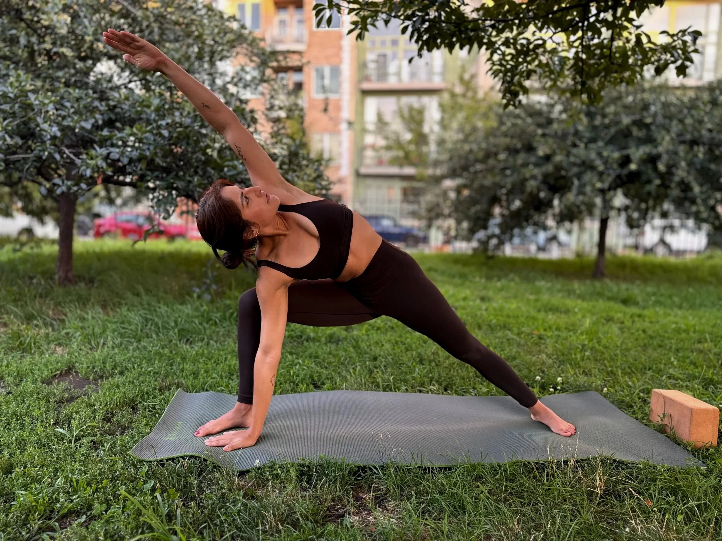 A woman practicing yoga outdoors on a mat in a park with trees and a city building in the background, performing a side angle pose with one arm extended upwards.