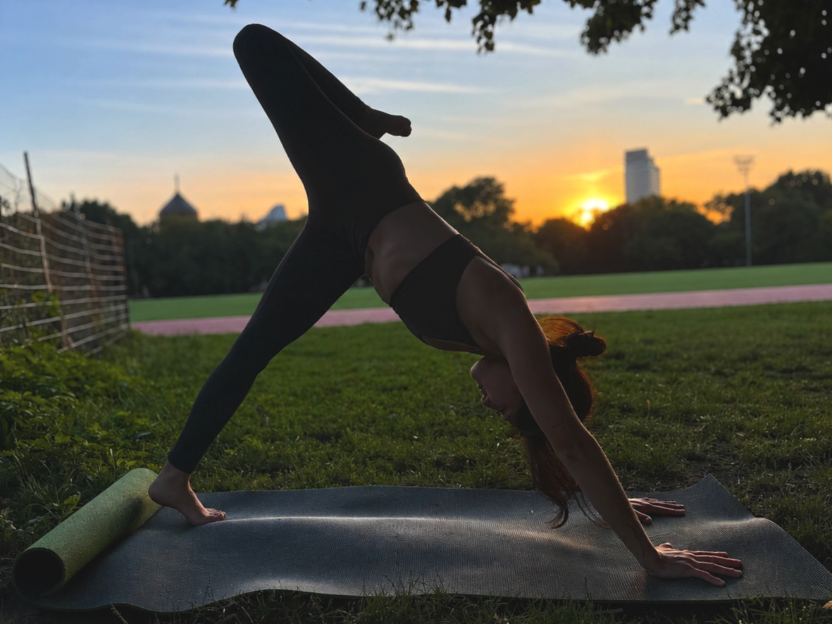 A woman practicing yoga outdoors on a mat during sunset, balancing in a downward dog pose with a sunset sky and cityscape in the background.
