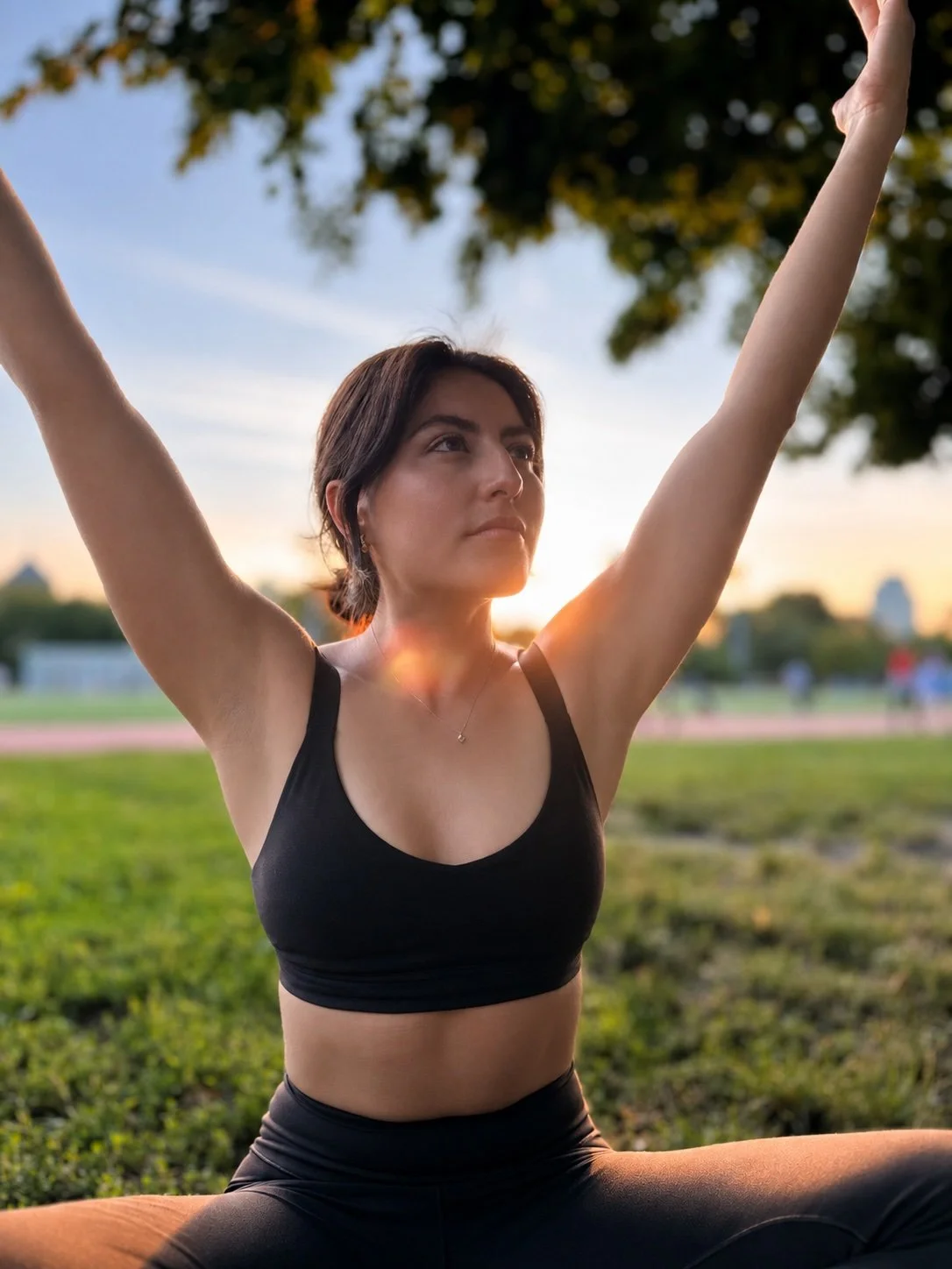 Woman practicing yoga outdoors at sunset, sitting cross-legged on the grass, with her arms raised overhead, wearing a brown sports bra and brown leggings.