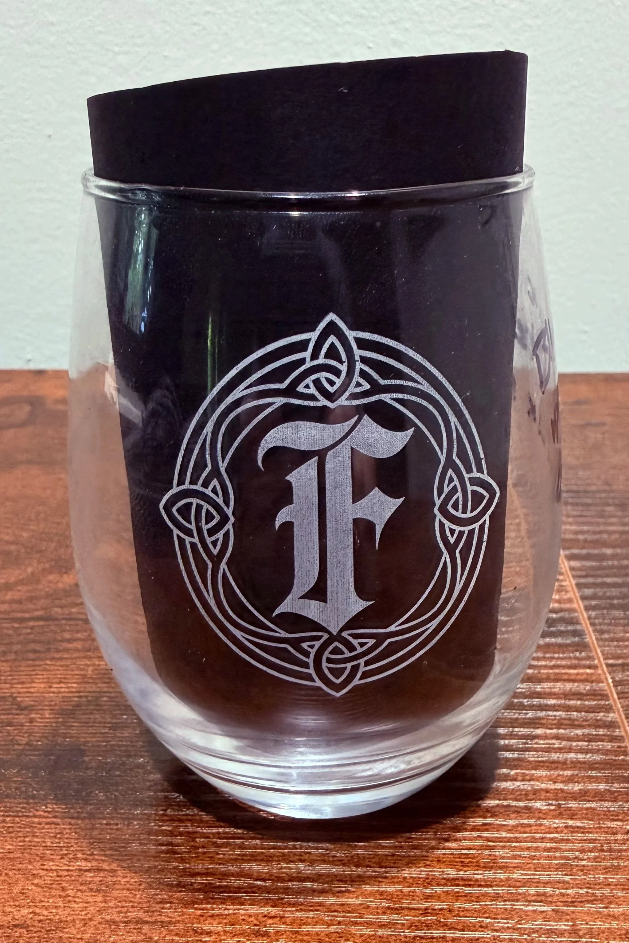 A stemless wine glass (black background to show frosted engraving), decorated with a white intricate Celtic knot design on the front, placed on a wooden surface against a light-colored wall.