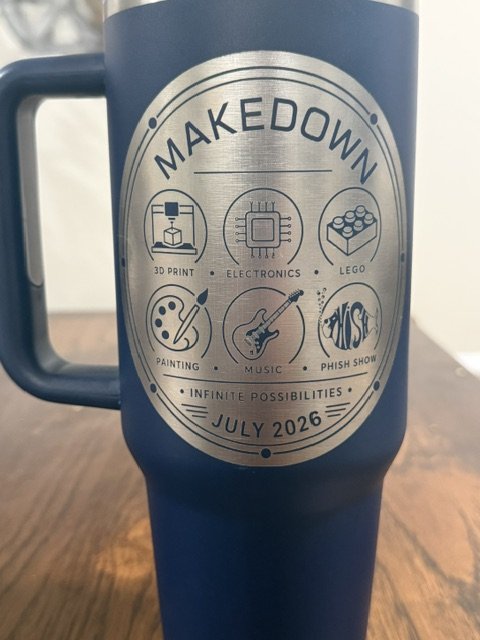 Blue mug with a circular design featuring icons and text for an event called Makedown, including 3D printing, electronics, Lego, painting, music, and a Phish show, with the date July 2026.