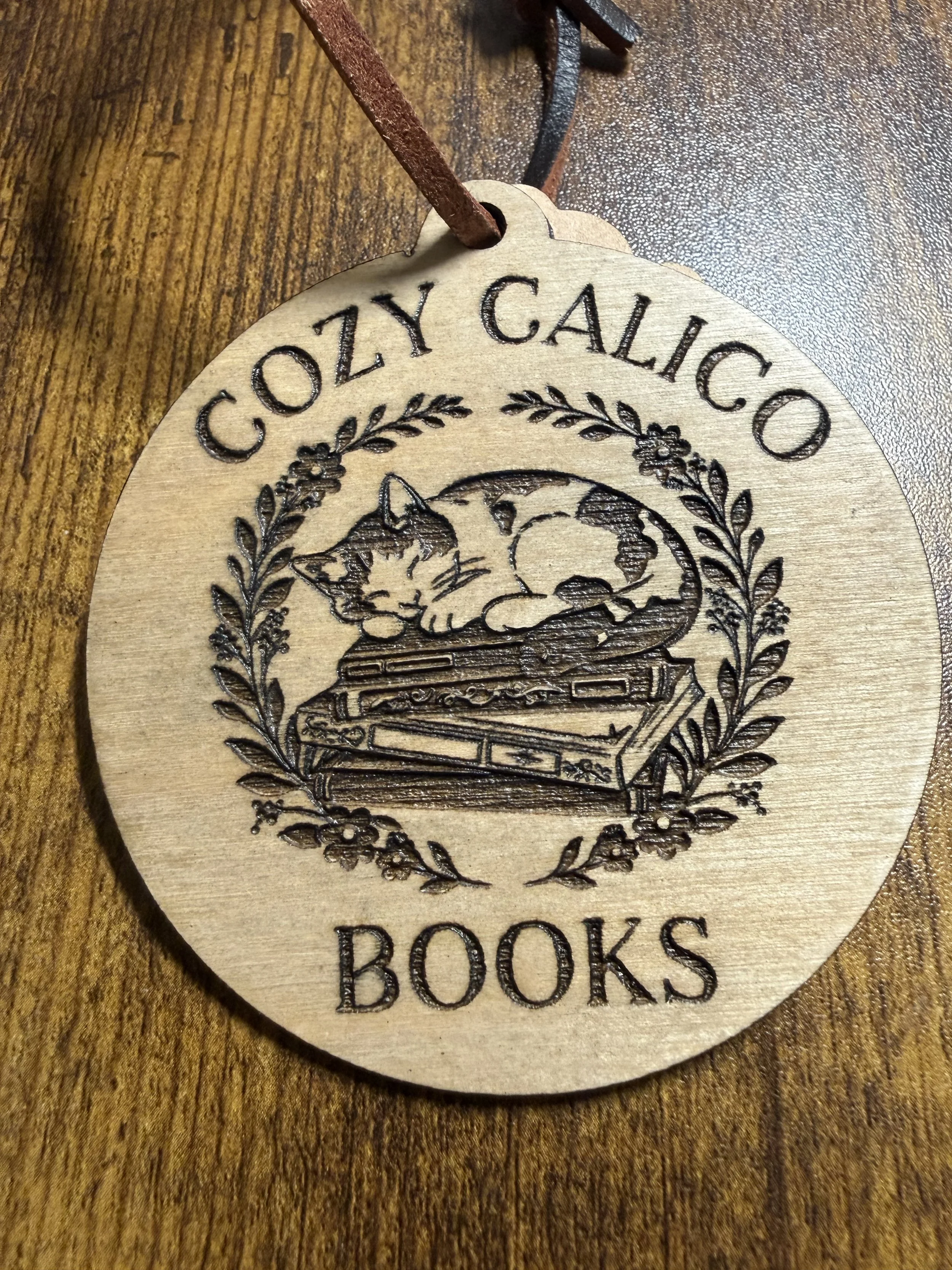A round wooden tag engraved with the words 'Cozy Calico' at the top and 'Books' at the bottom, featuring a detailed illustration of a cat curled up on a stack of books, surrounded by a wreath of flowers and leaves.