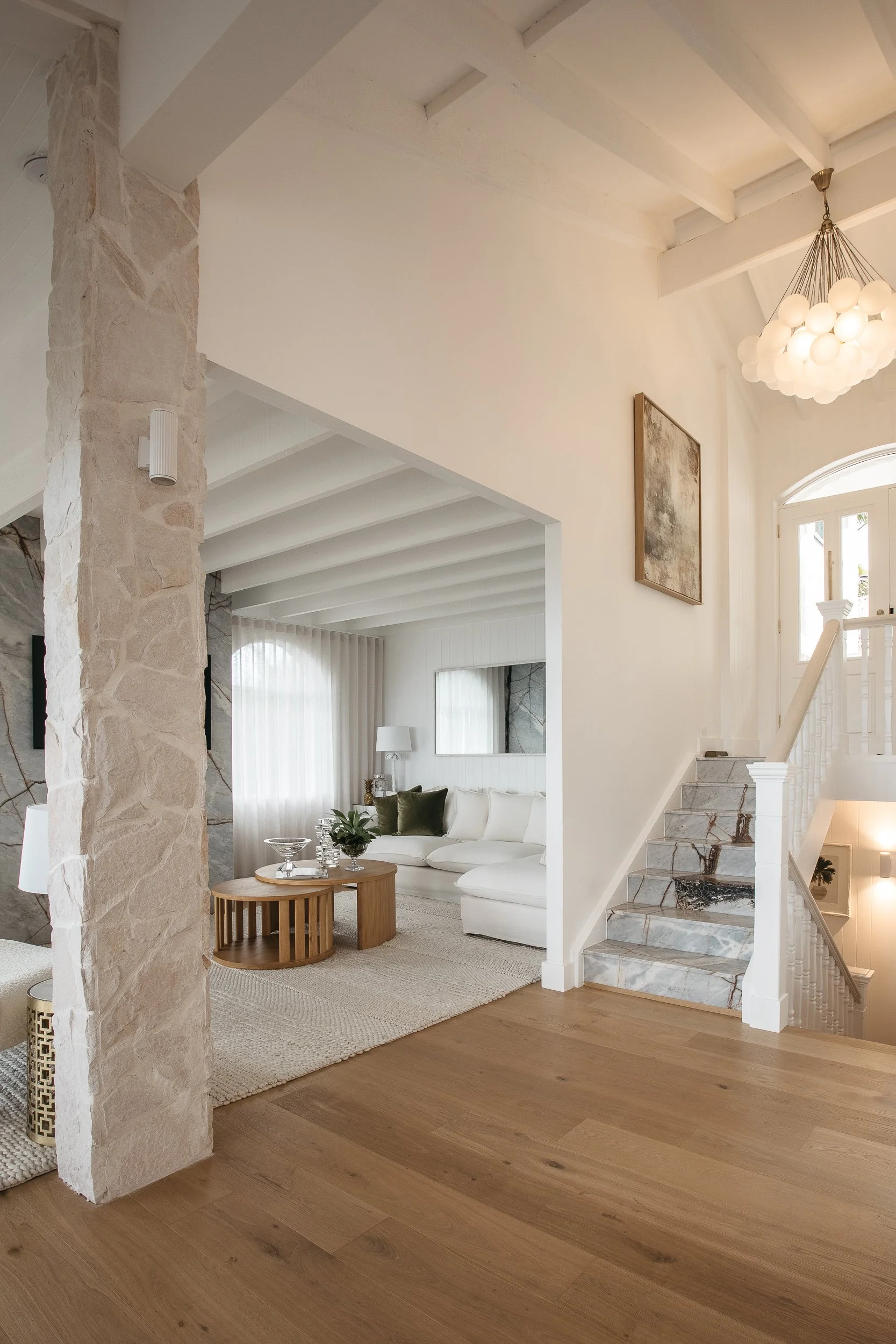 Living room with white walls, stone pillar, wooden flooring, and staircase with marble steps. A white sofa with green and white pillows, round wooden coffee table with plants and decorative items, area rug, framed artwork on wall, and a chandelier hanging from the ceiling.