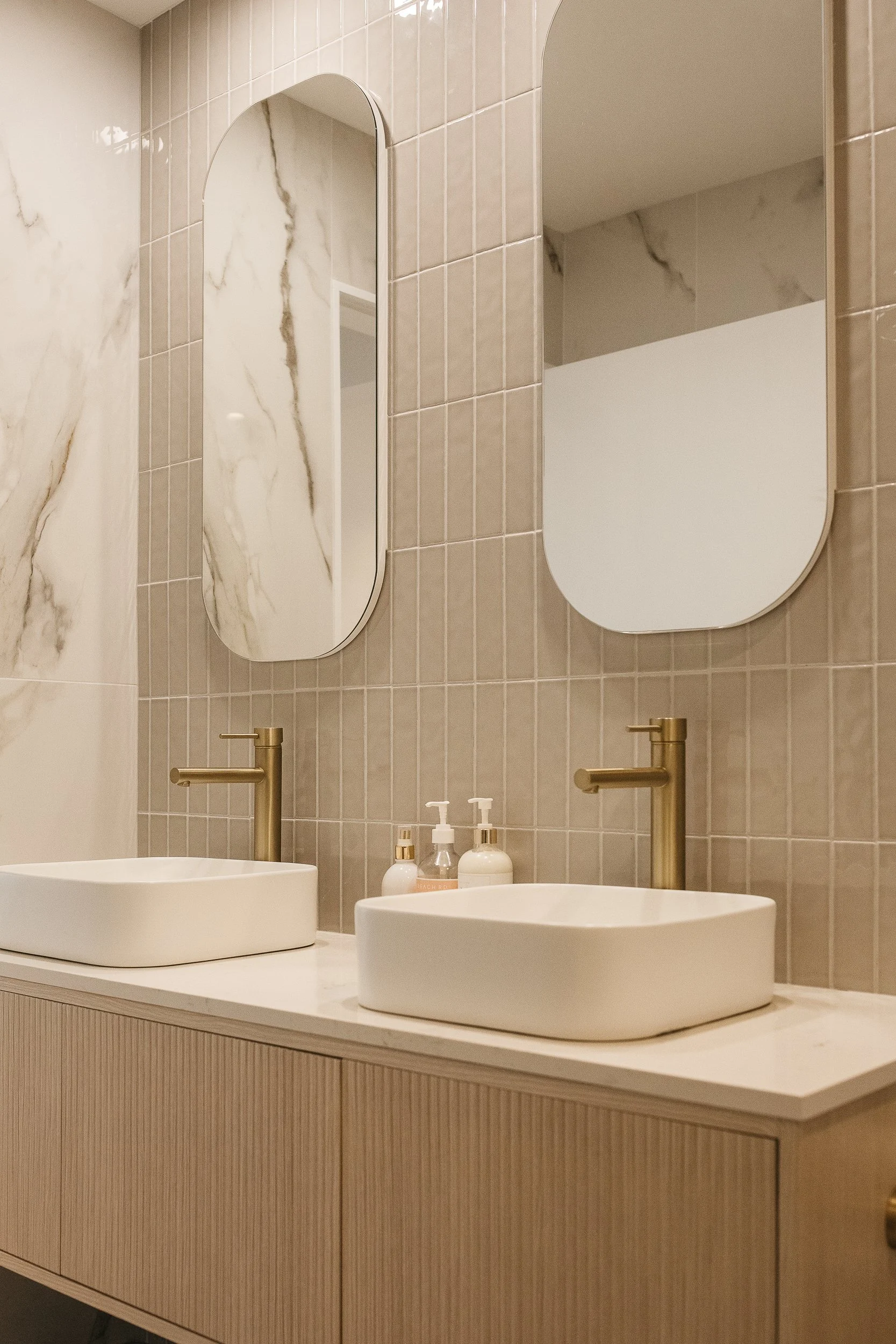 Modern bathroom vanity with two white vessel sinks, brushed gold faucets, and two large oval mirrors on beige tiled wall, with soap dispensers on countertop.