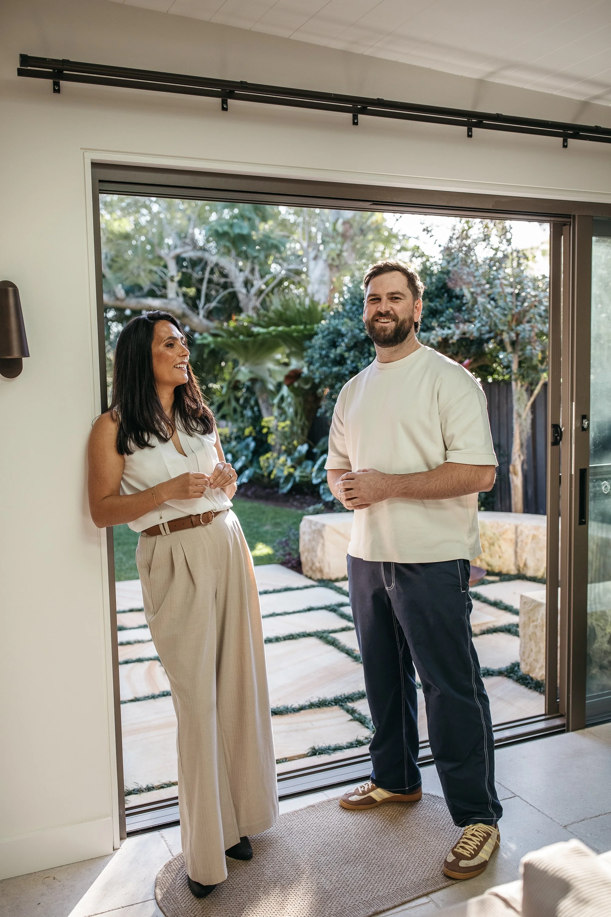 A woman and a man standing in a doorway, smiling and talking to each other, with a garden visible outside.