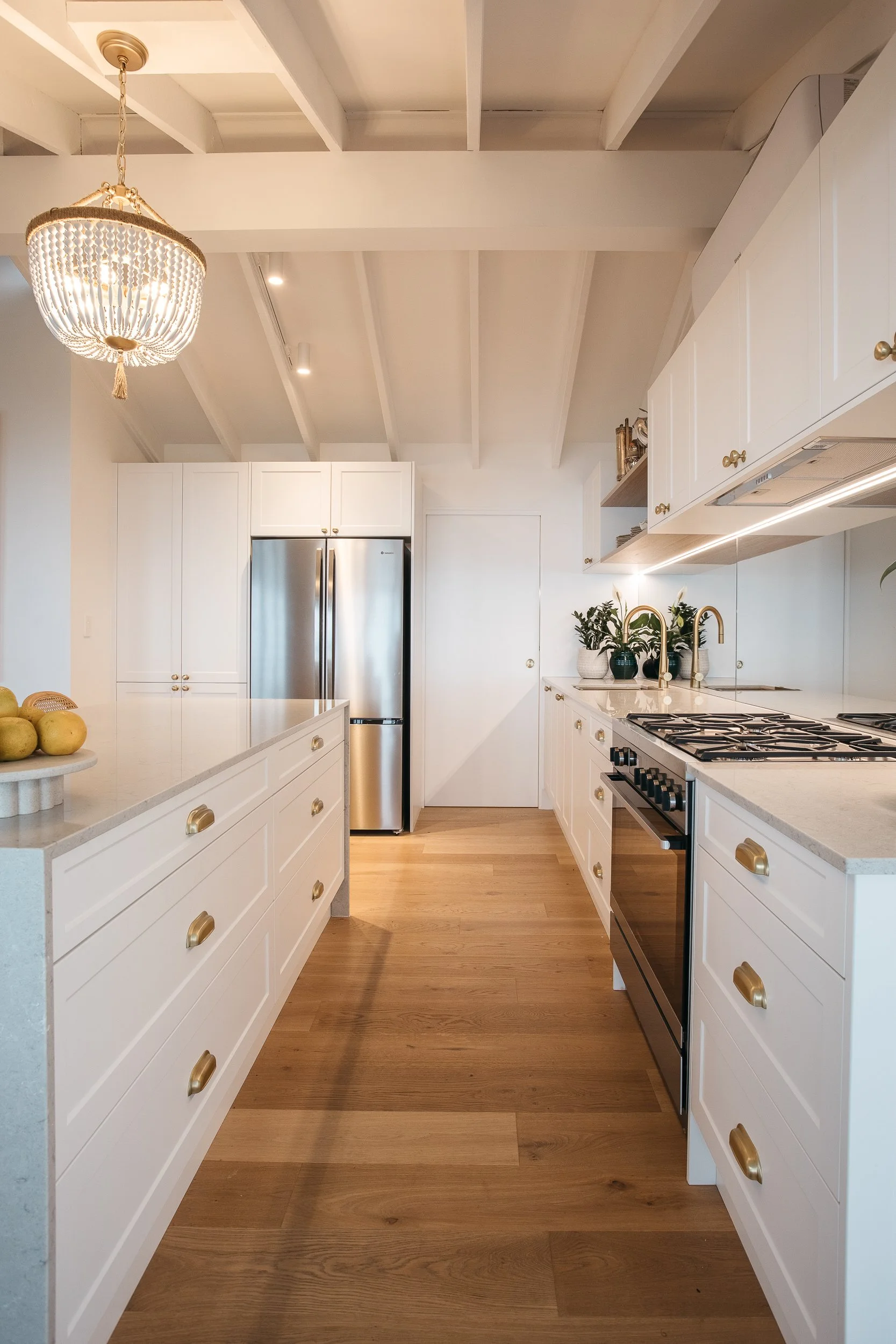 A modern kitchen with white cabinets, gold handles, a stainless steel refrigerator, a gas oven, wooden flooring, and decorative plants on the counter. There is a large chandelier hanging from the ceiling.