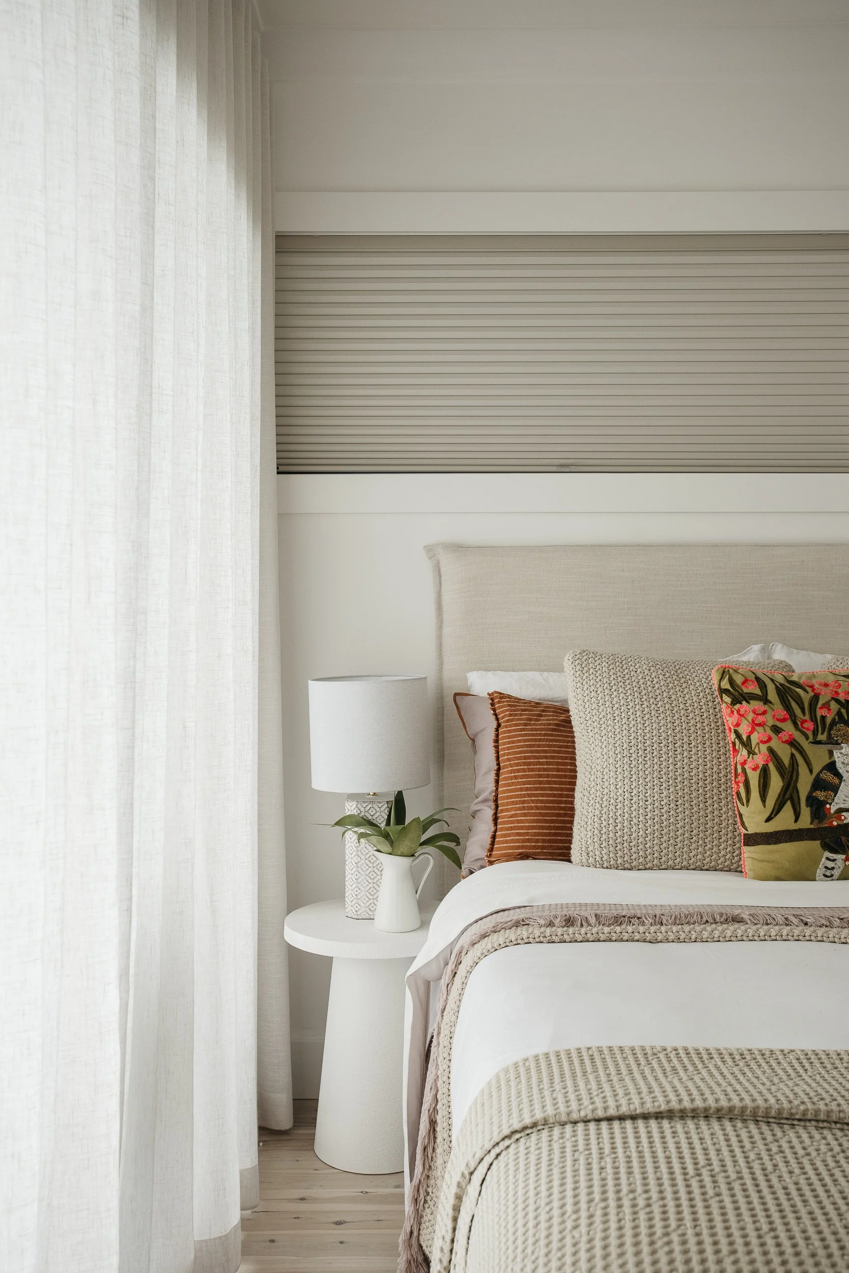 A bedroom corner with a bed that has a beige upholstered headboard and decorative pillows in various colors and patterns, next to a white side table with a potted plant and a white table lamp. Light-colored curtains partially cover a window behind the bed.