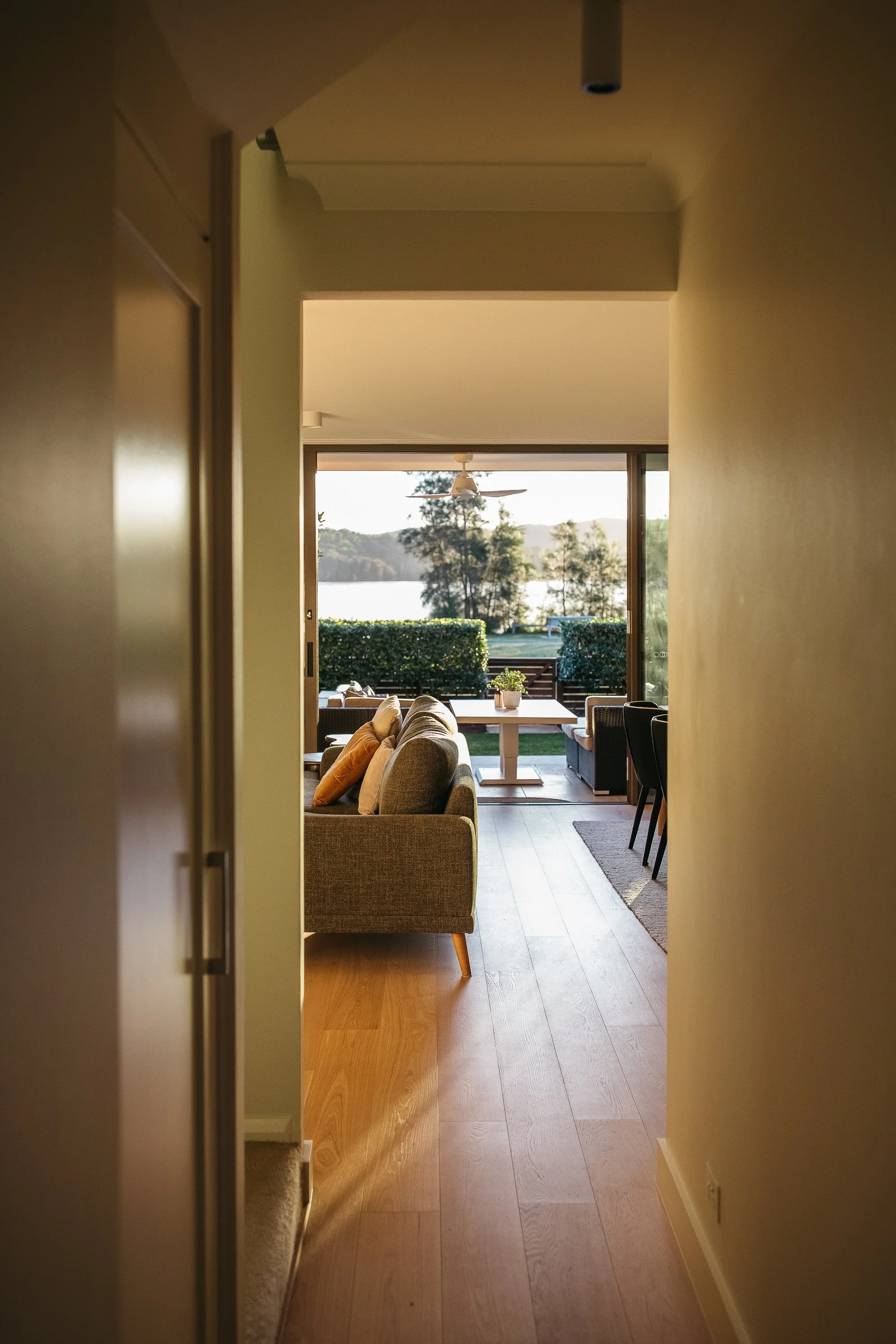 View through a doorway into a living room with a couch, chairs, a dining table, and a large window showing trees and water outside.
