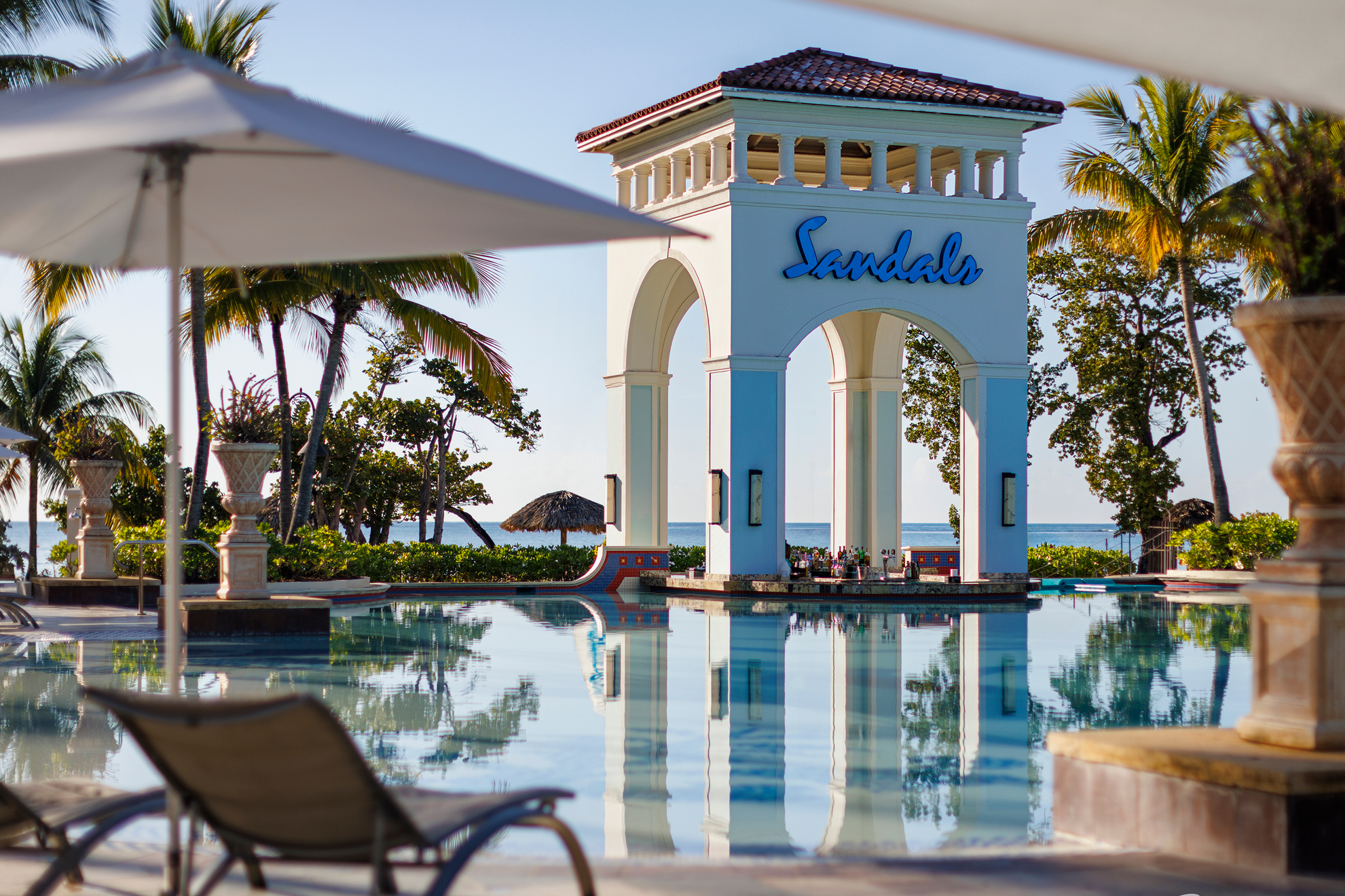 Resort pool area with lounge chairs, umbrellas, and a large archway structure with the word 'Sandals' on it, surrounded by palm trees and tropical plants.
