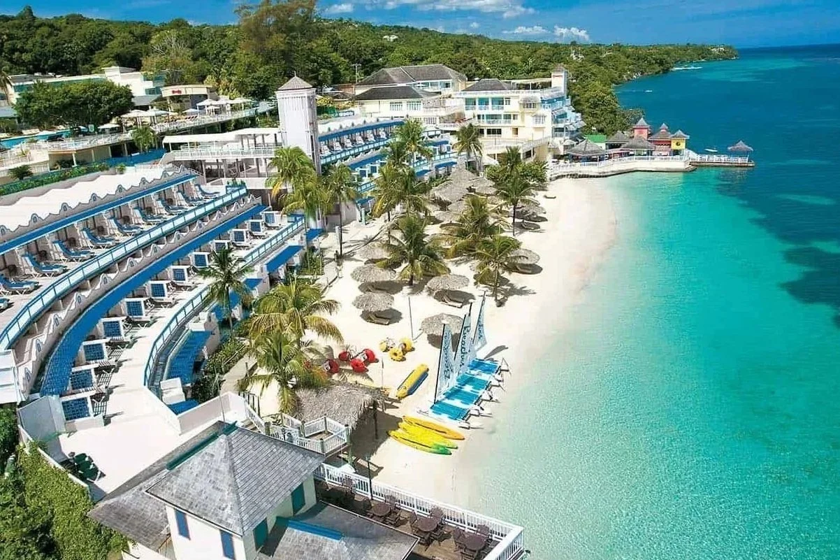 Aerial view of a beach resort with white sandy beach, turquoise water, palm trees, umbrellas, and a multi-story hotel building with balconies overlooking the water.