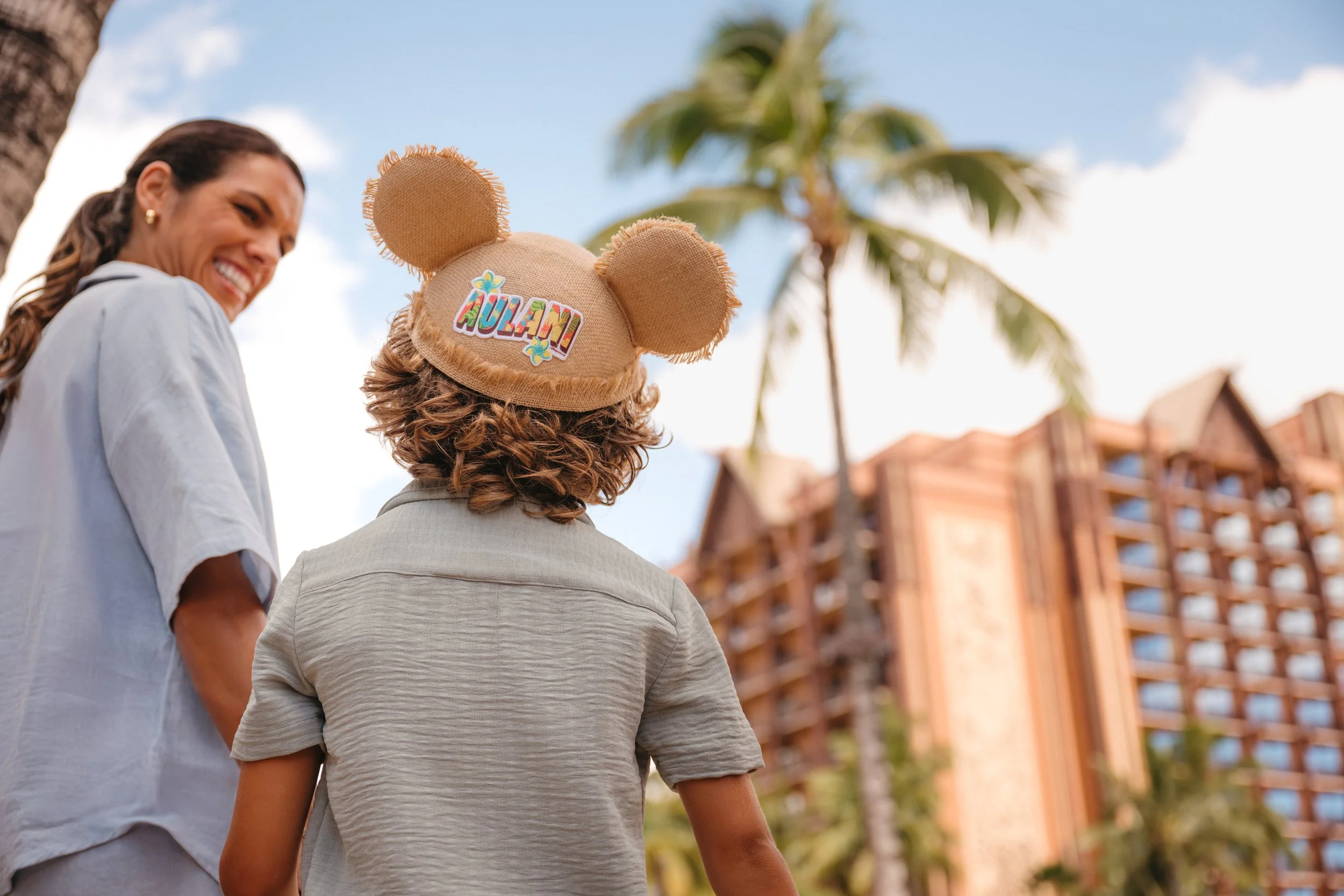 A smiling woman in a white top walking outdoors with another person wearing a beige hat with Mickey ears and a colorful sticker that says "AULANI". Palm trees and a building are in the background.