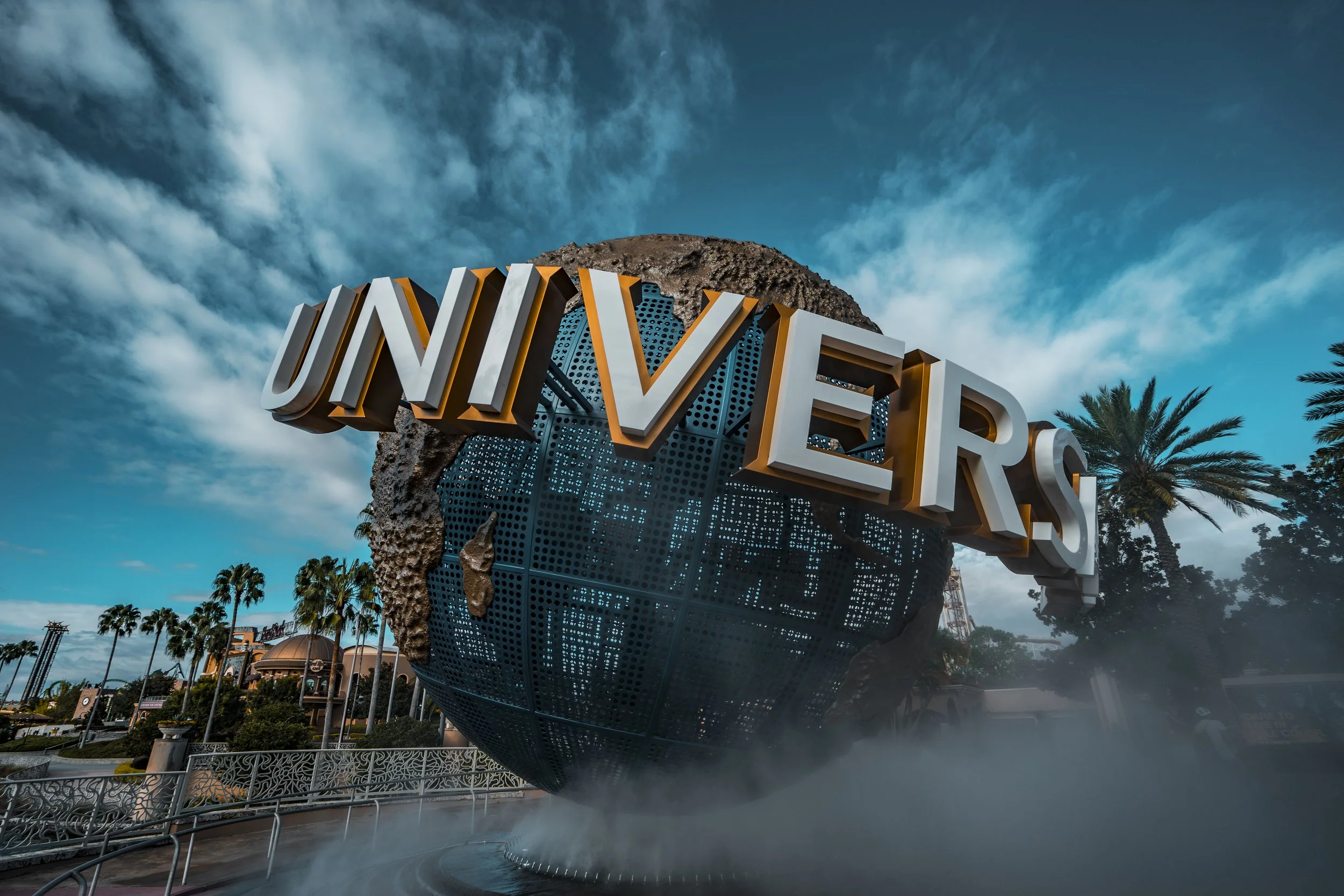 A large sign with the word 'UNIVERSAL' hangs atop a globe sculpture outside Universal Studios theme park, with palm trees and a partly cloudy sky in the background.