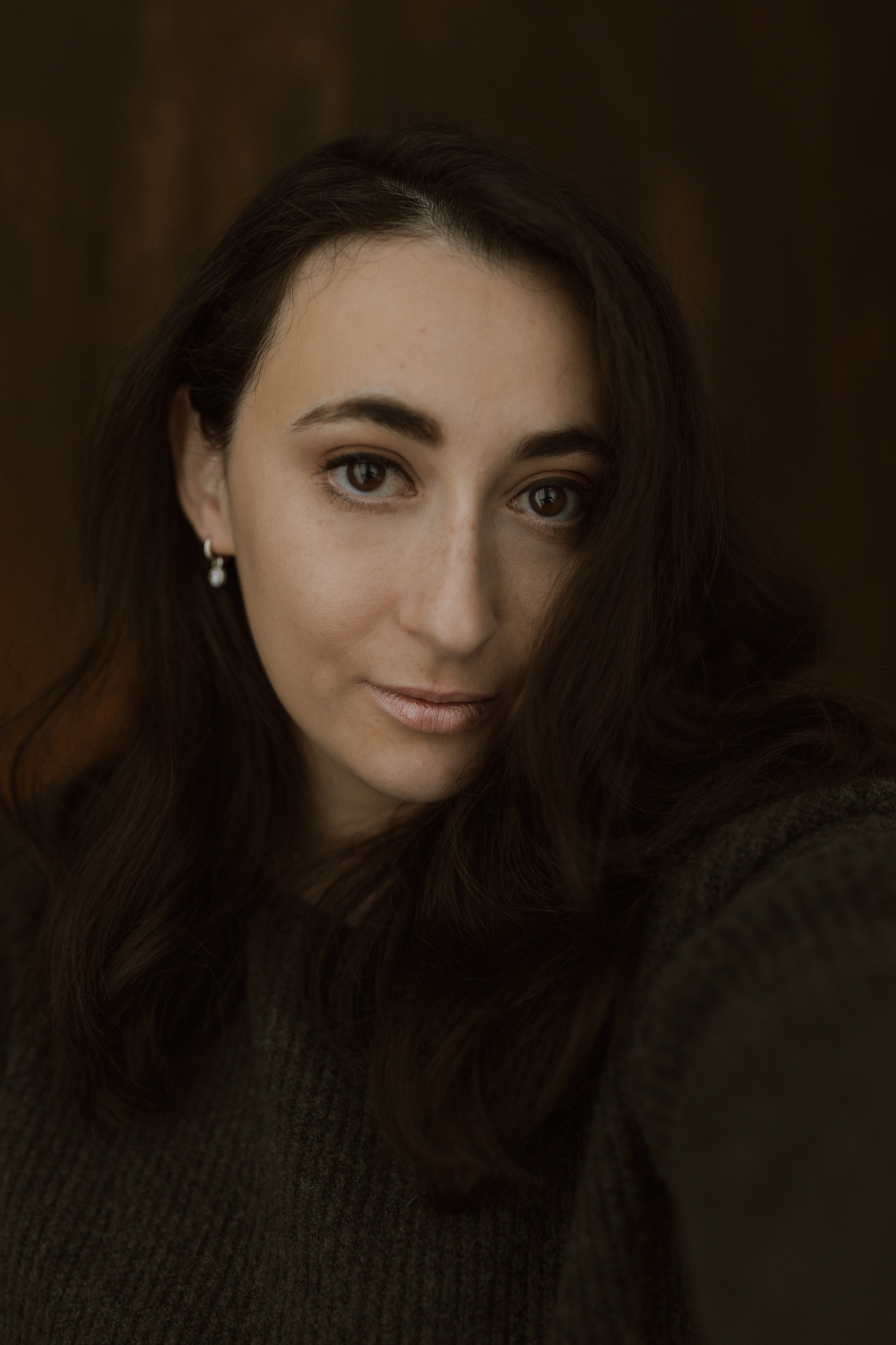 Close-up portrait of a woman with long, wavy brown hair, light skin, brown eyes, wearing earrings and a dark sweater.